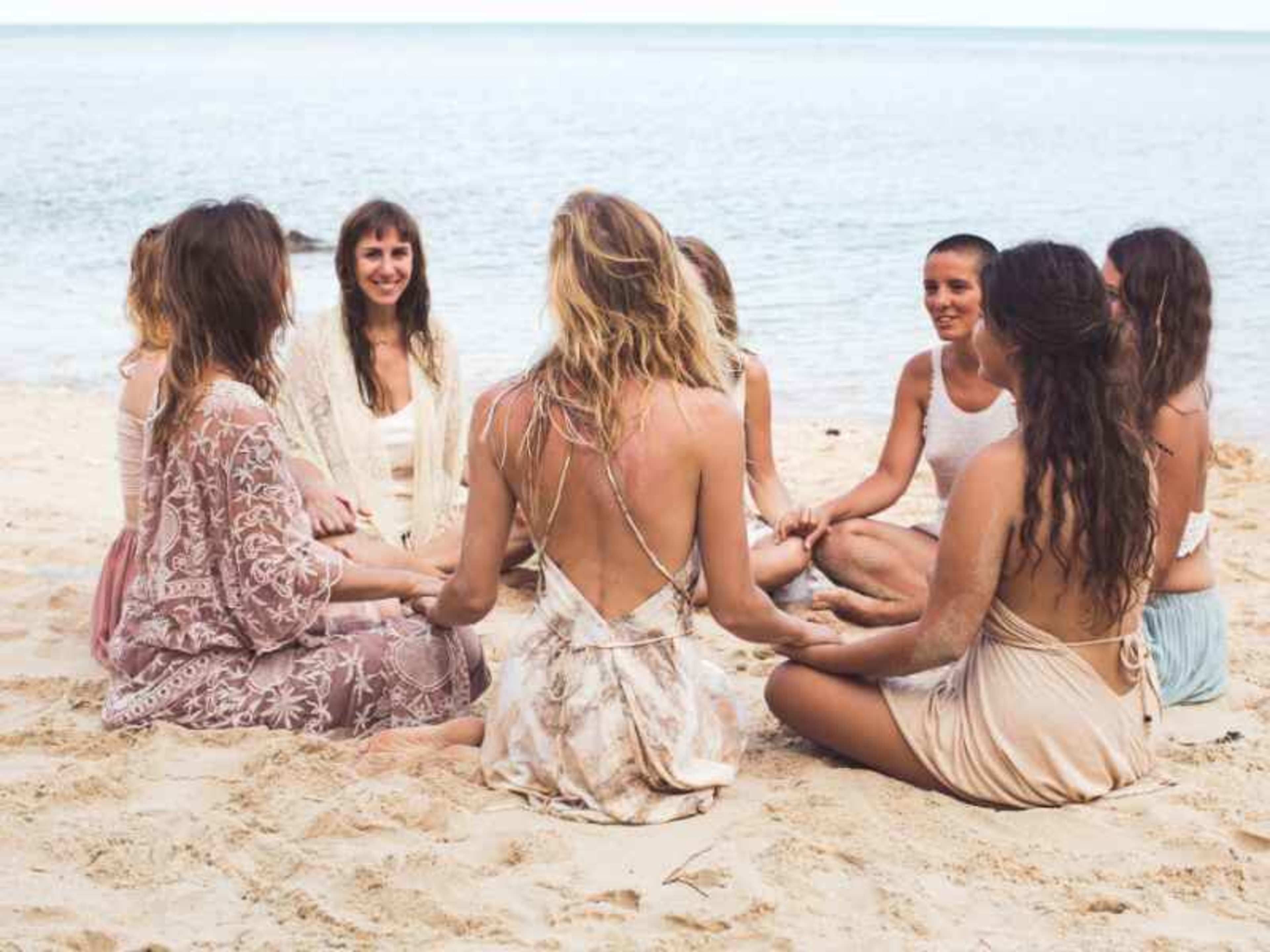 A group of women in light clothing sits in a circle on the beach, facing each other near the water.