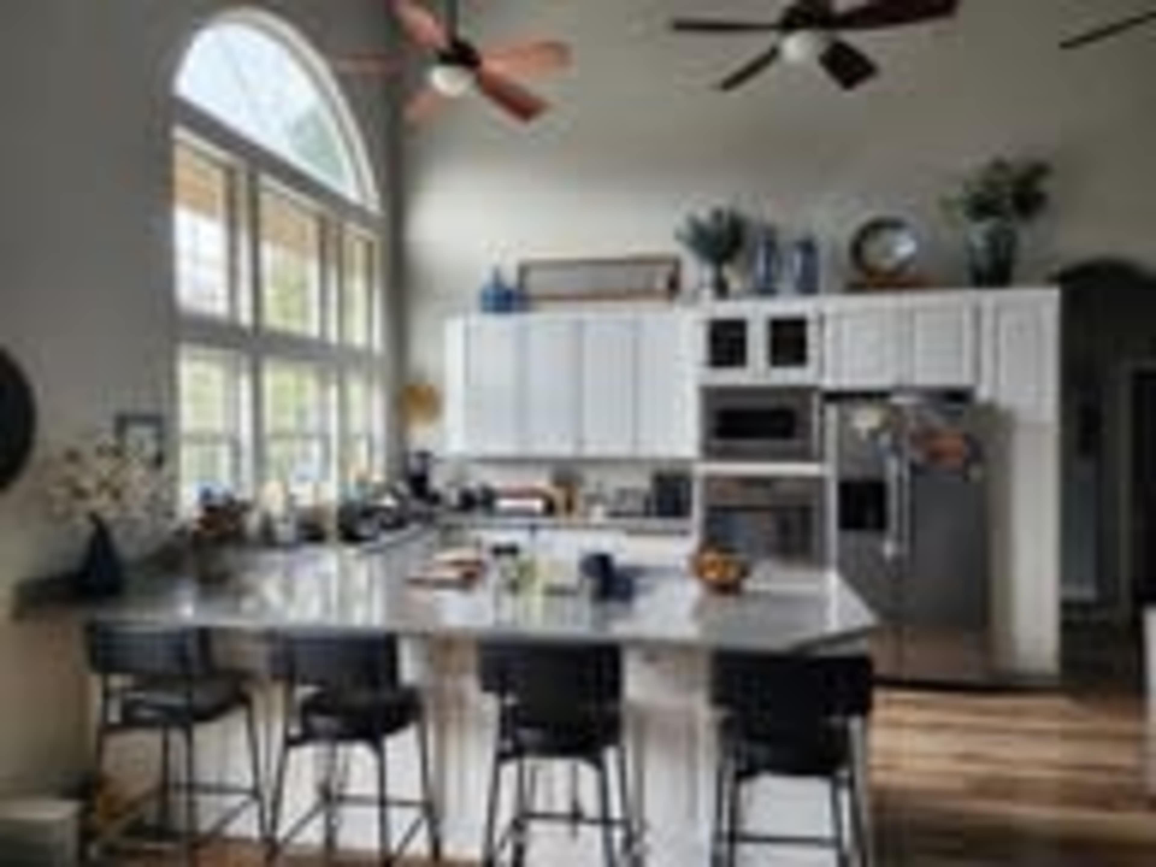 A modern kitchen features white cabinetry, stainless steel appliances, a large island with seating, and abundant natural light streaming through arched windows.