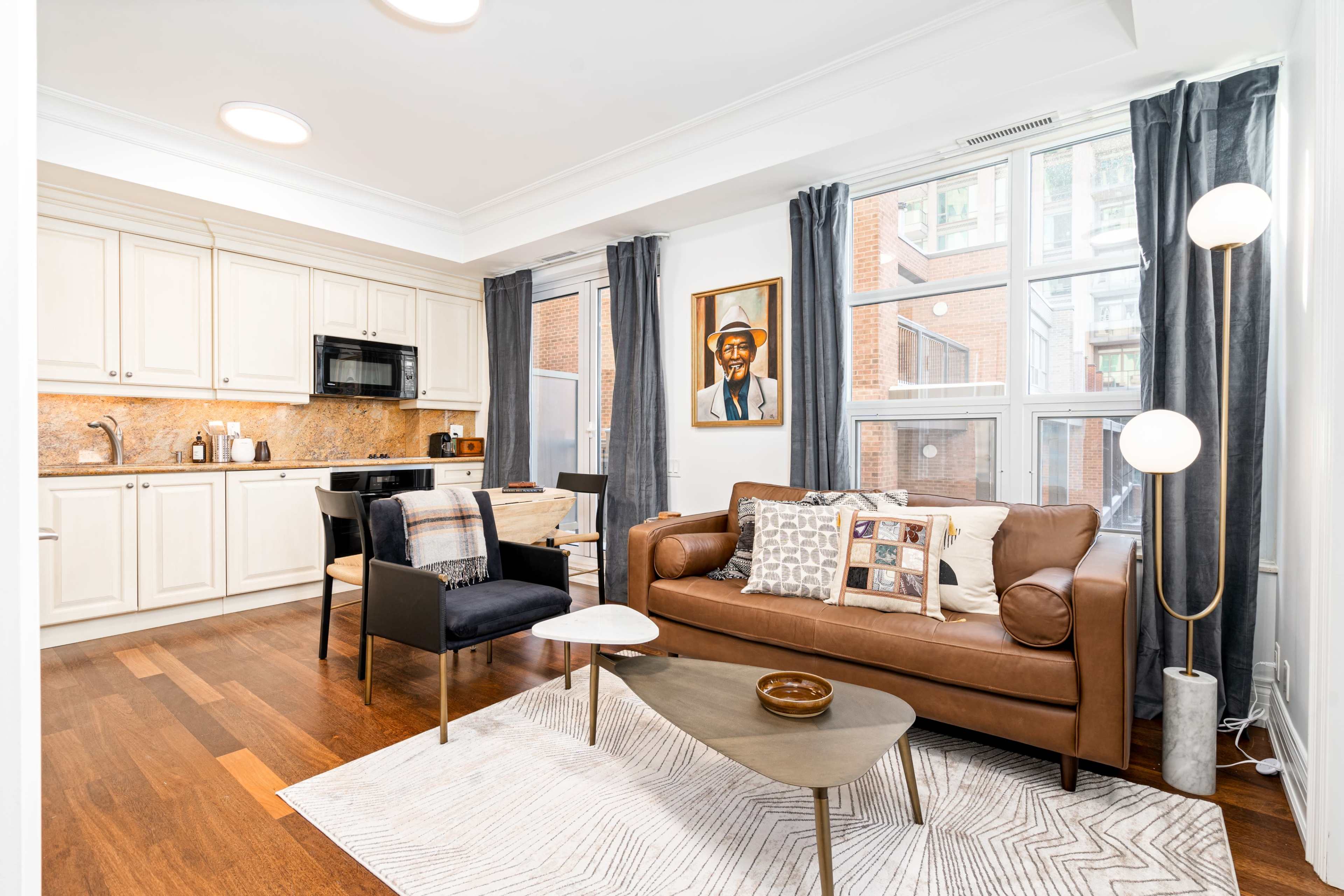 A modern apartment living area featuring a kitchen with light cabinetry, a sitting area with a brown leather couch and a small coffee table, and large windows providing natural light.