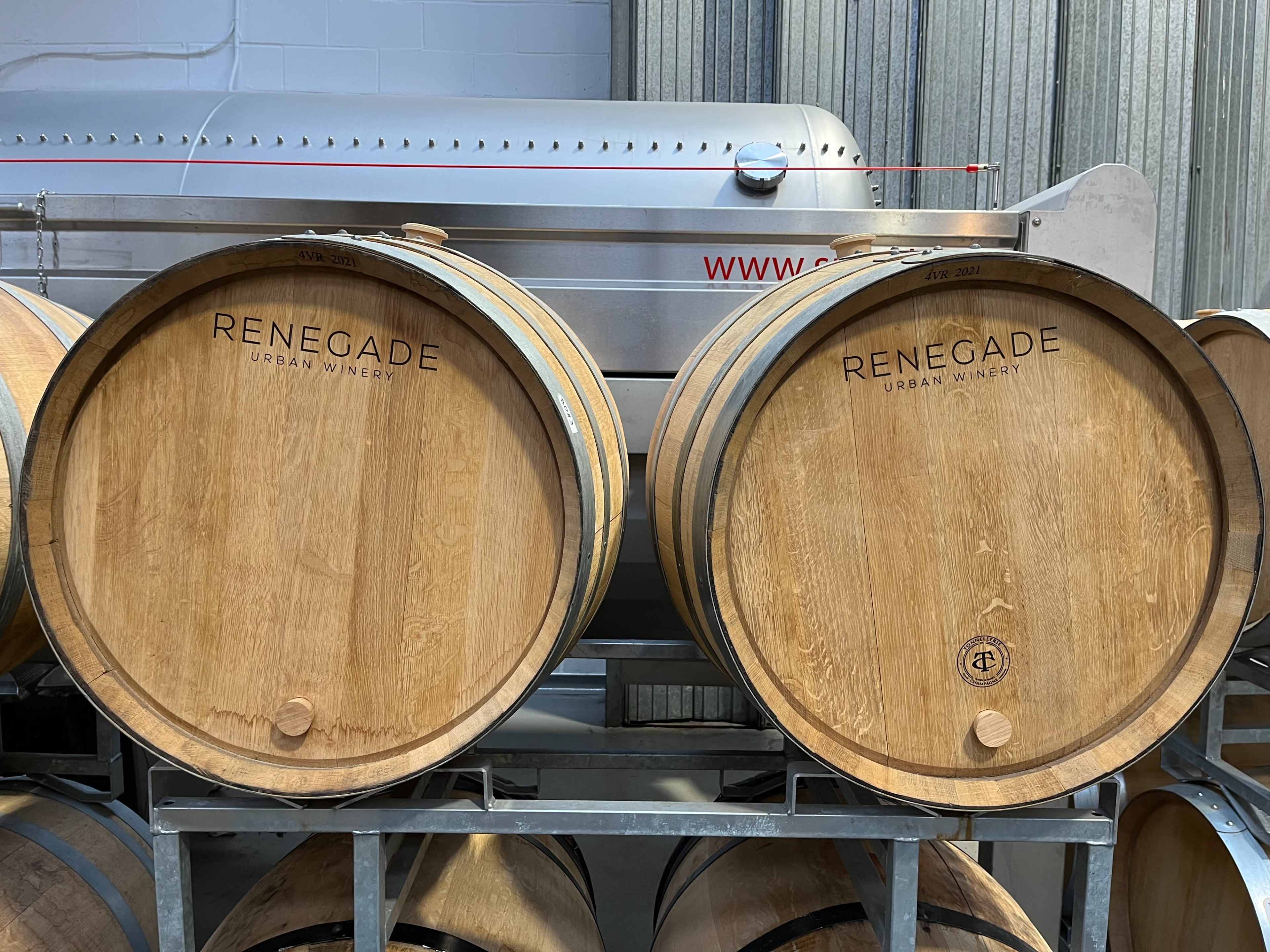 Two wooden wine barrels labeled with "RENEGADE URBAN WINERY" are stacked on a metal rack in a winery setting.