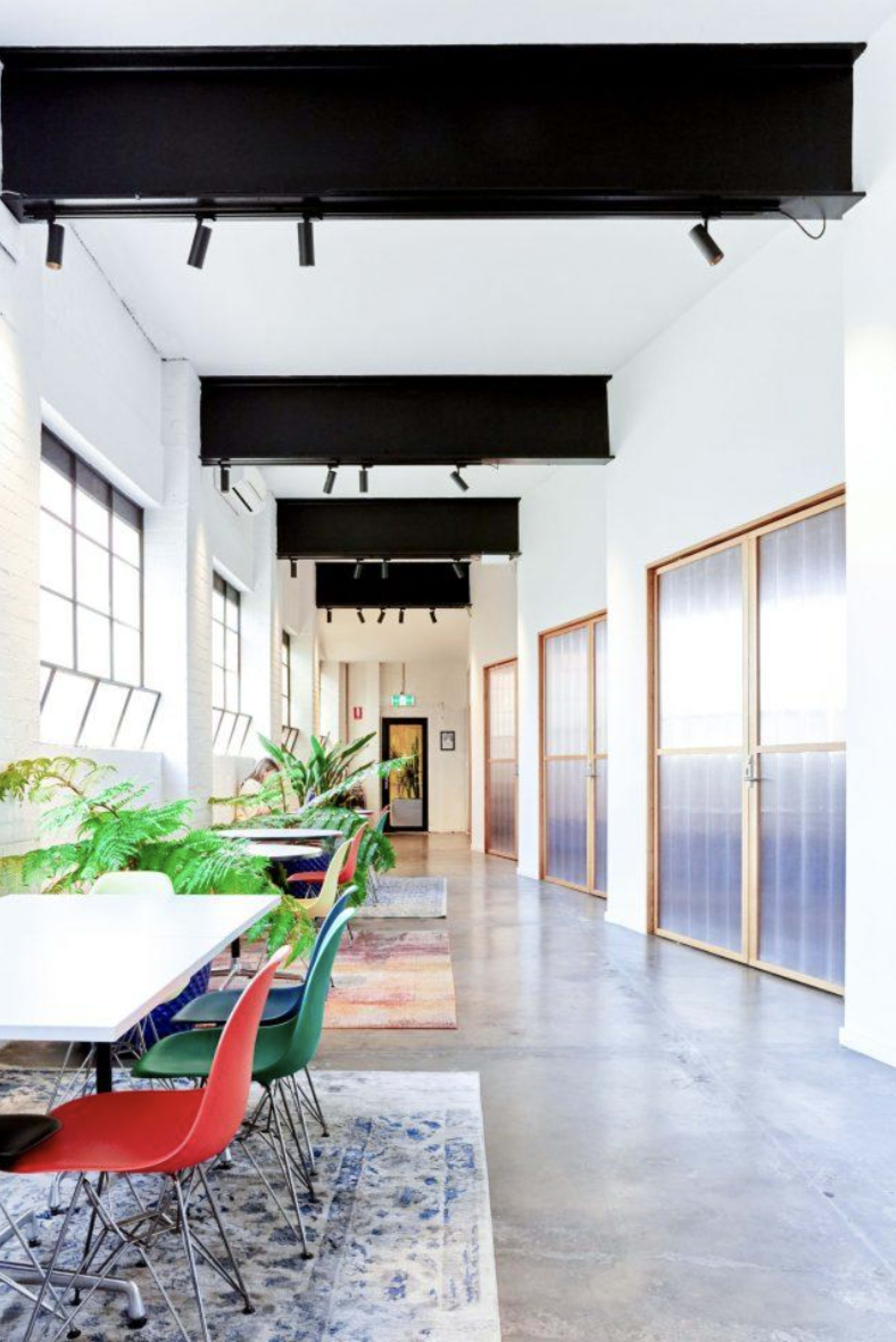 A modern hallway with a polished concrete floor, lined with plants and colorful chairs around a white table, leading to glass-paneled doors.