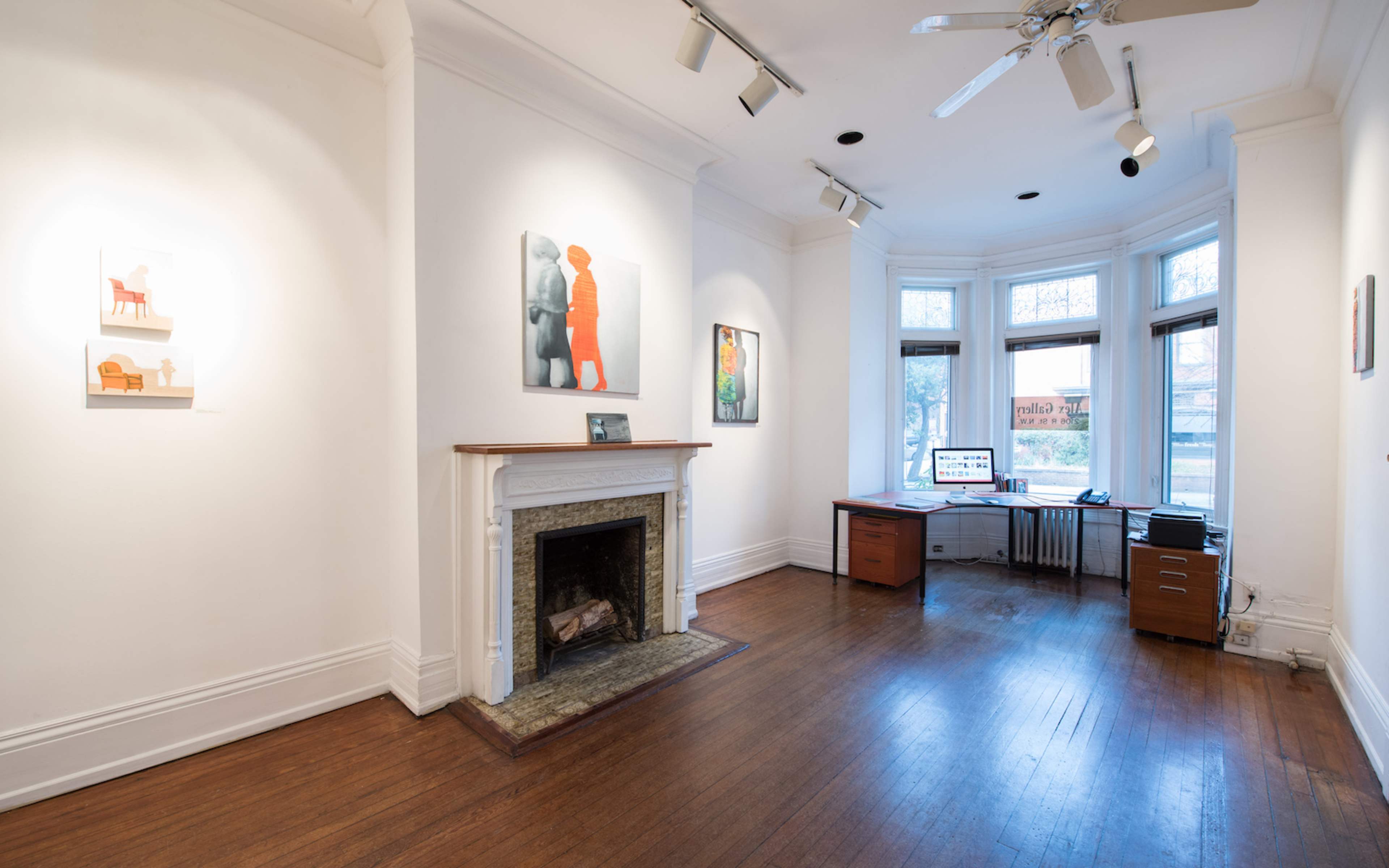 A spacious room with white walls, a fireplace, a wooden floor, and a desk with a computer positioned near large windows.