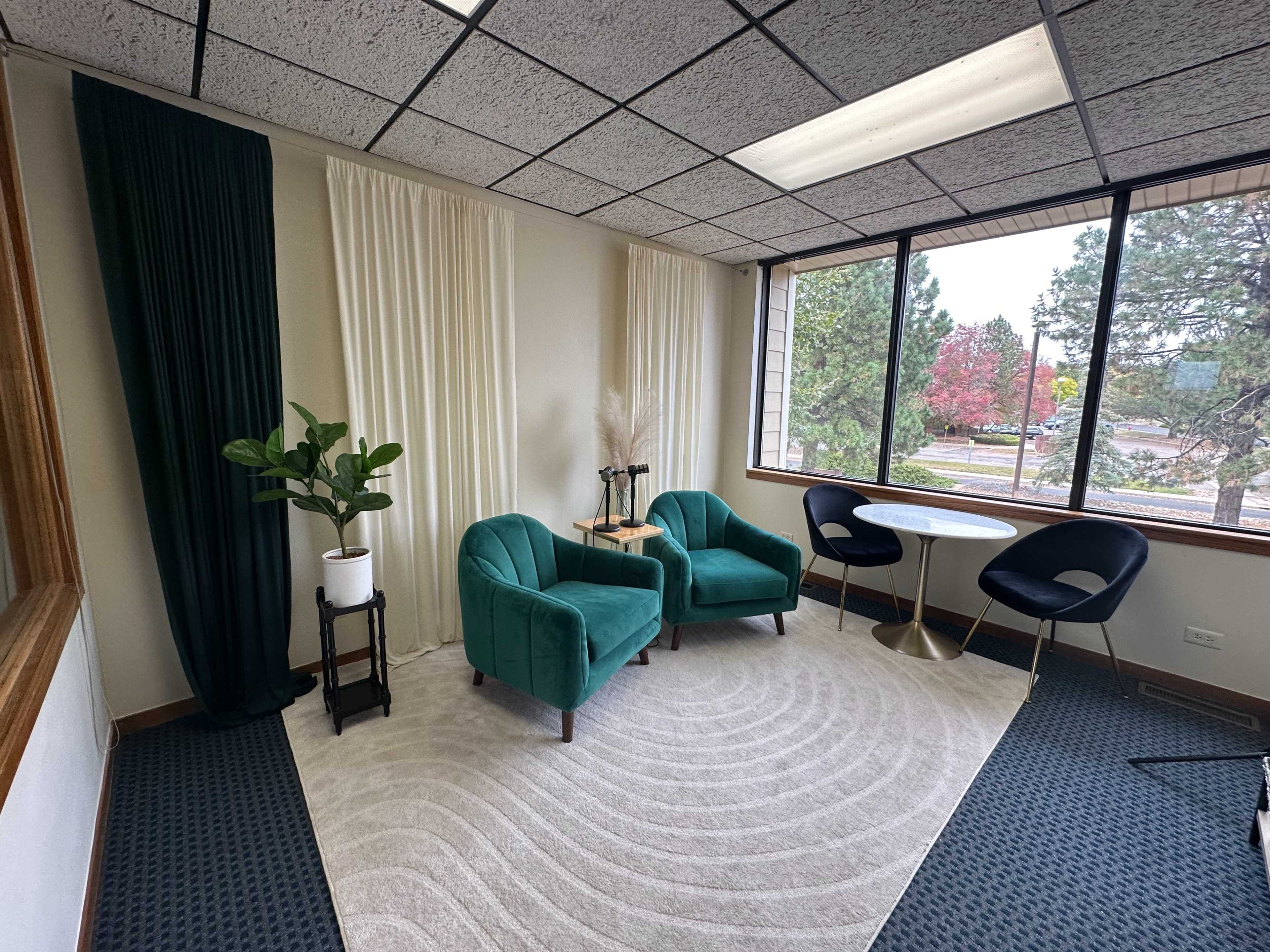 The image shows a cozy office space featuring two teal chairs, a small round table, and a potted plant, with large windows revealing a view of trees outside.
