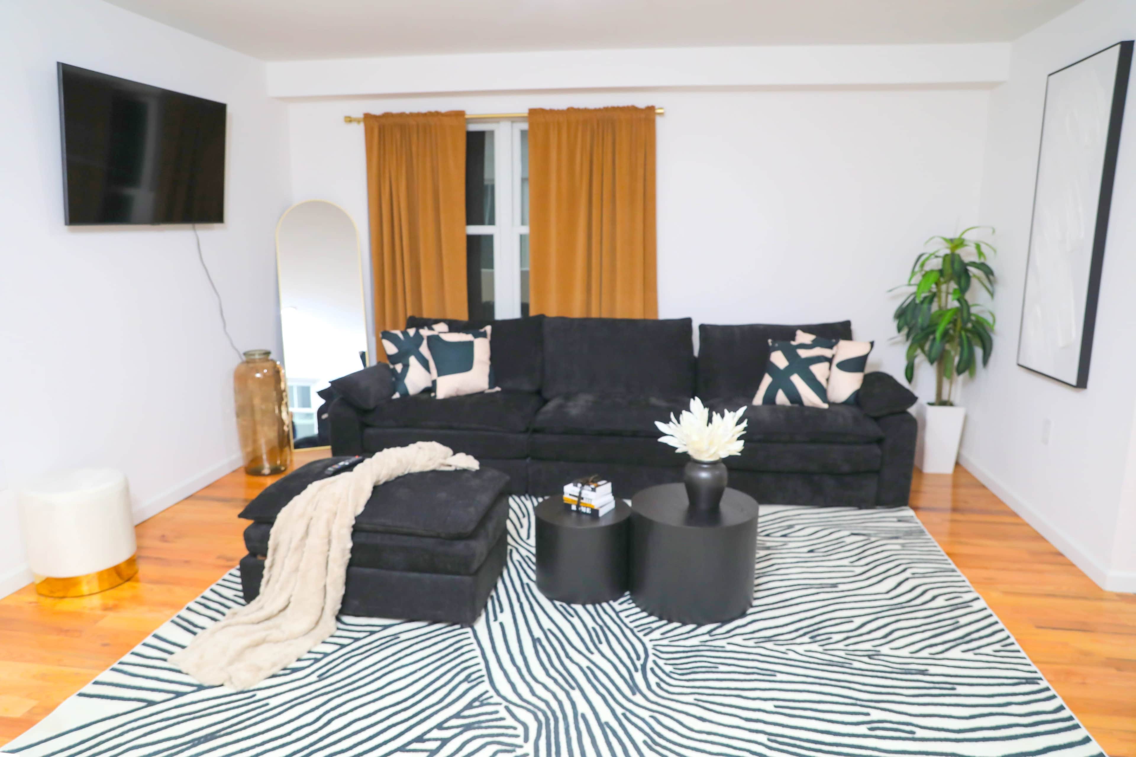The living room features a black sofa with decorative pillows, a circular black coffee table, and a zebra-striped rug, accompanied by a large mirror and a potted plant.