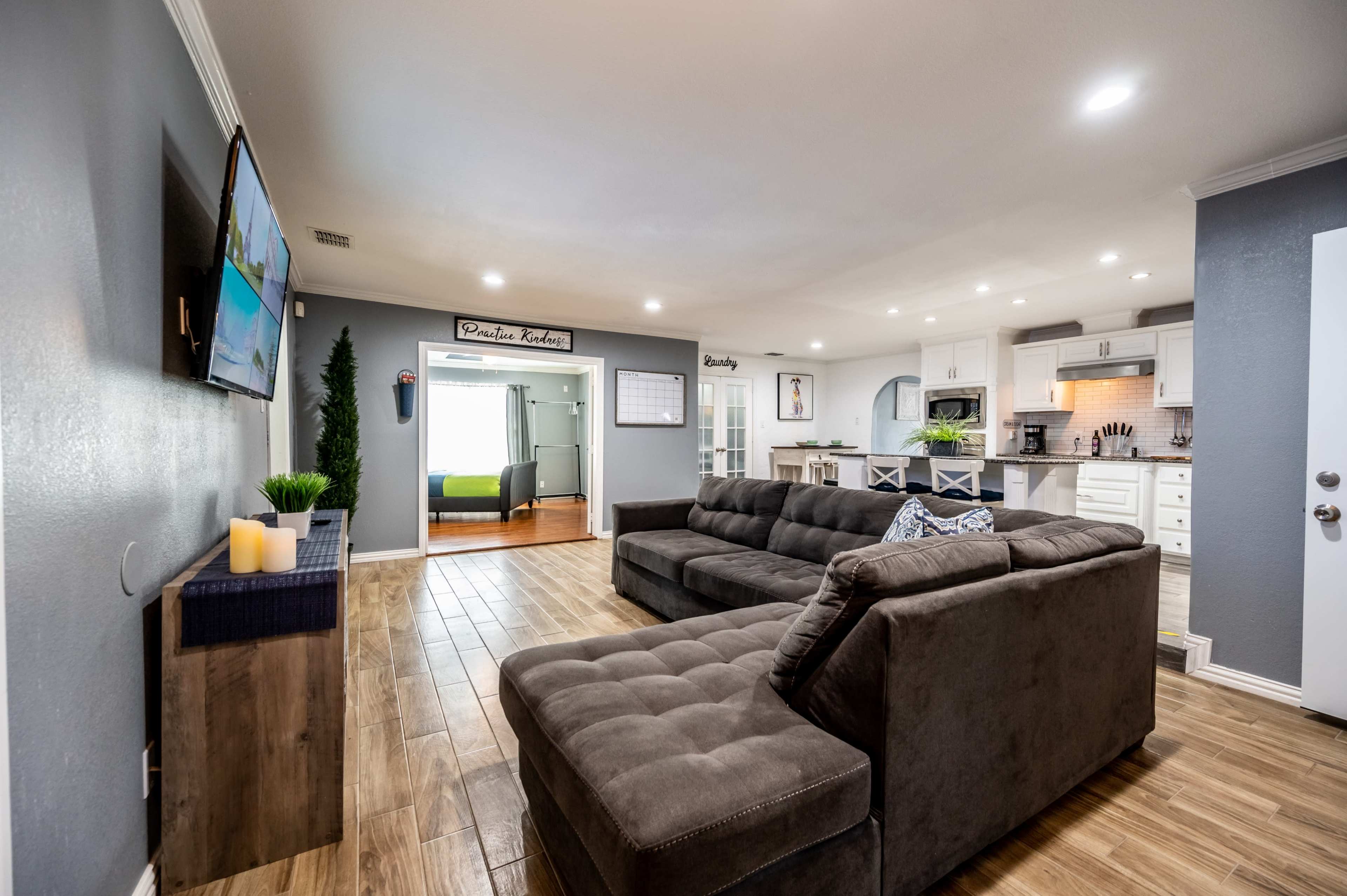 The image shows a modern living room with a large sectional sofa, a television mounted on the wall, and an open view into a kitchen area.