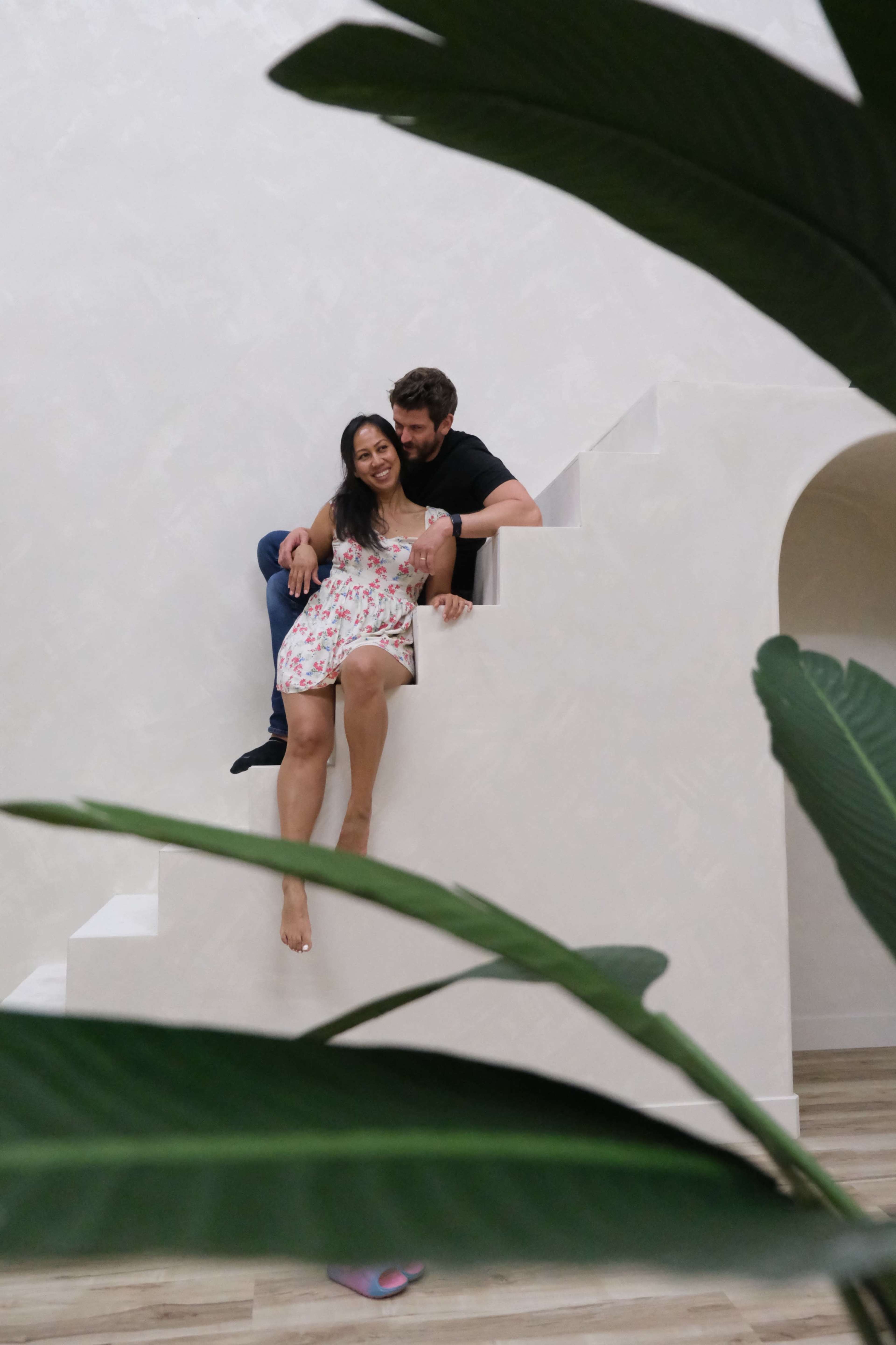 A couple sits on a white staircase while surrounded by green plants.