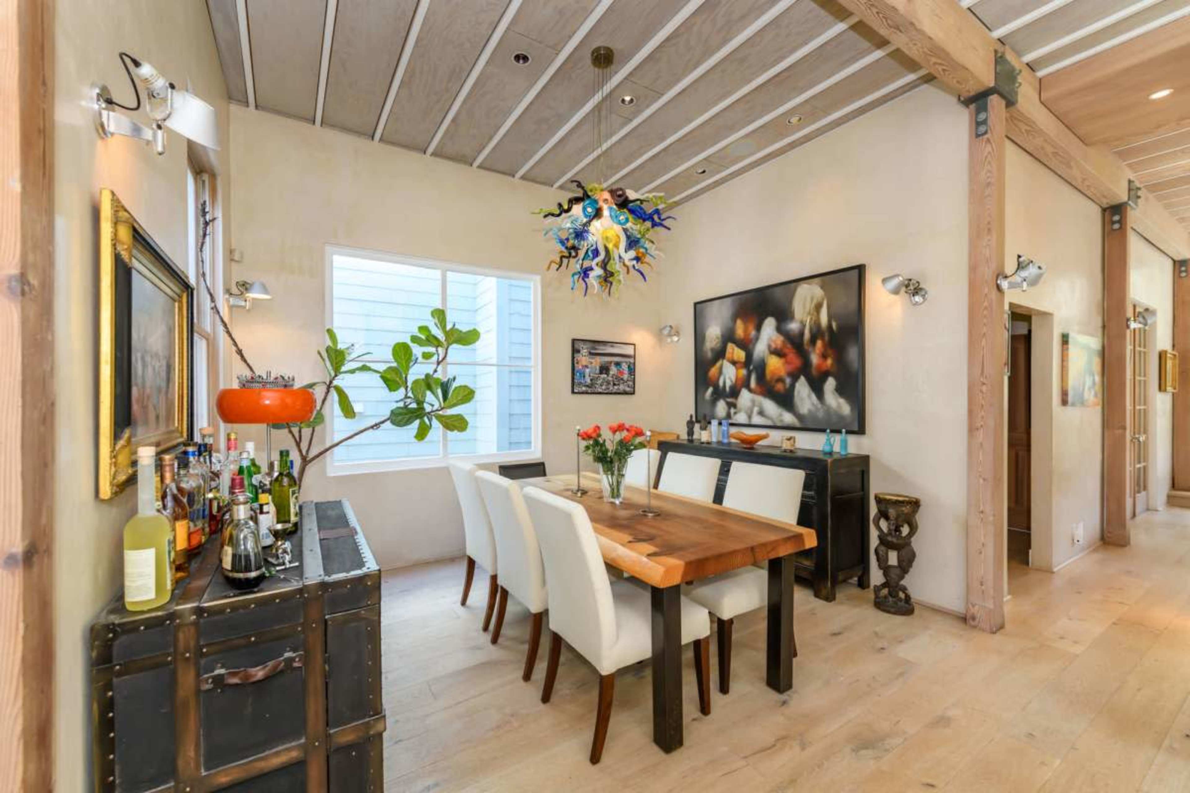 The image shows a dining area with a wooden table surrounded by white chairs, an organized bar cart, and artwork on the walls, all set in a bright, contemporary space.