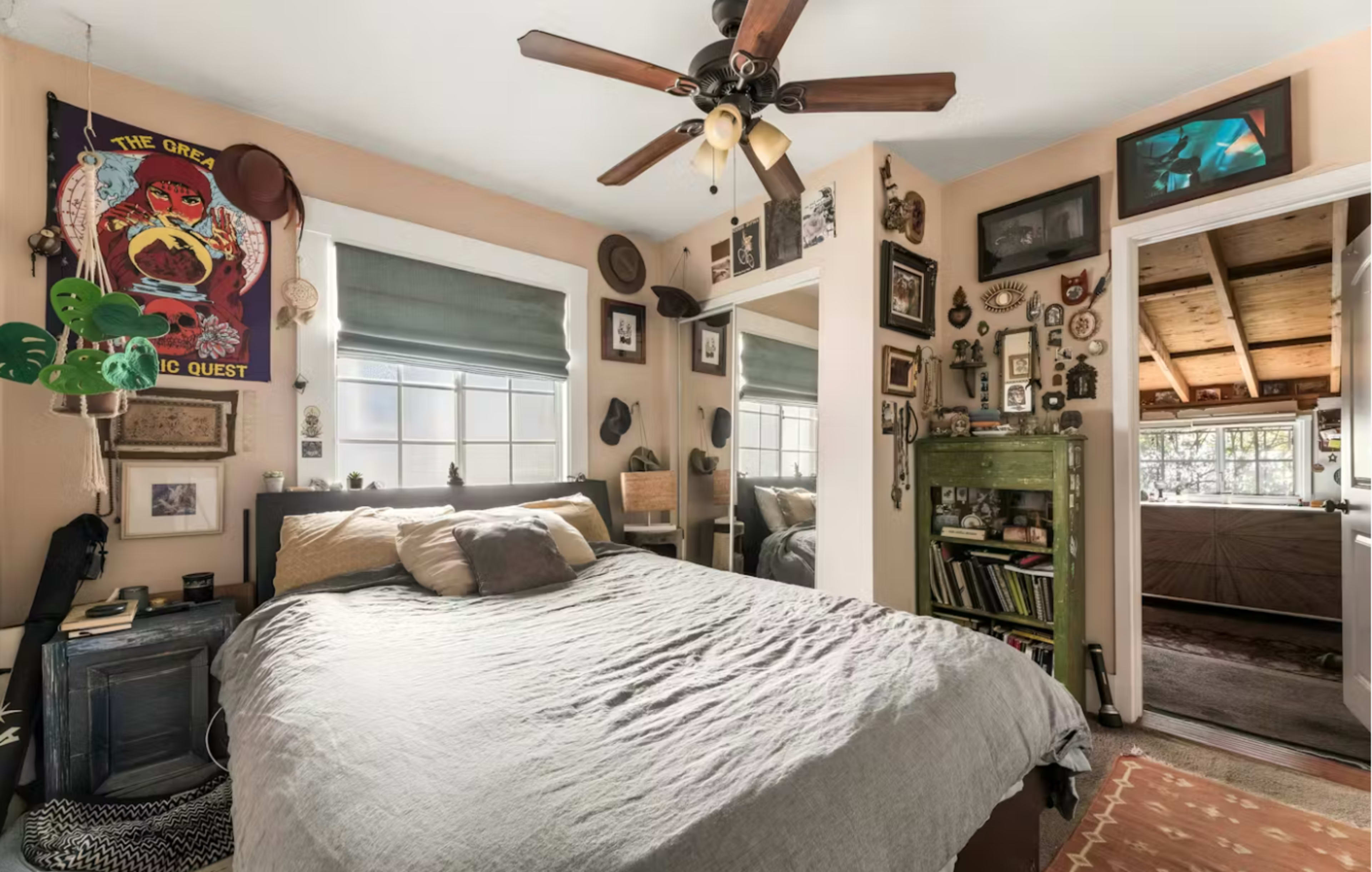 The image shows a cozy bedroom featuring a bed with gray bedding, a ceiling fan, and walls adorned with various framed pictures and decorative items.