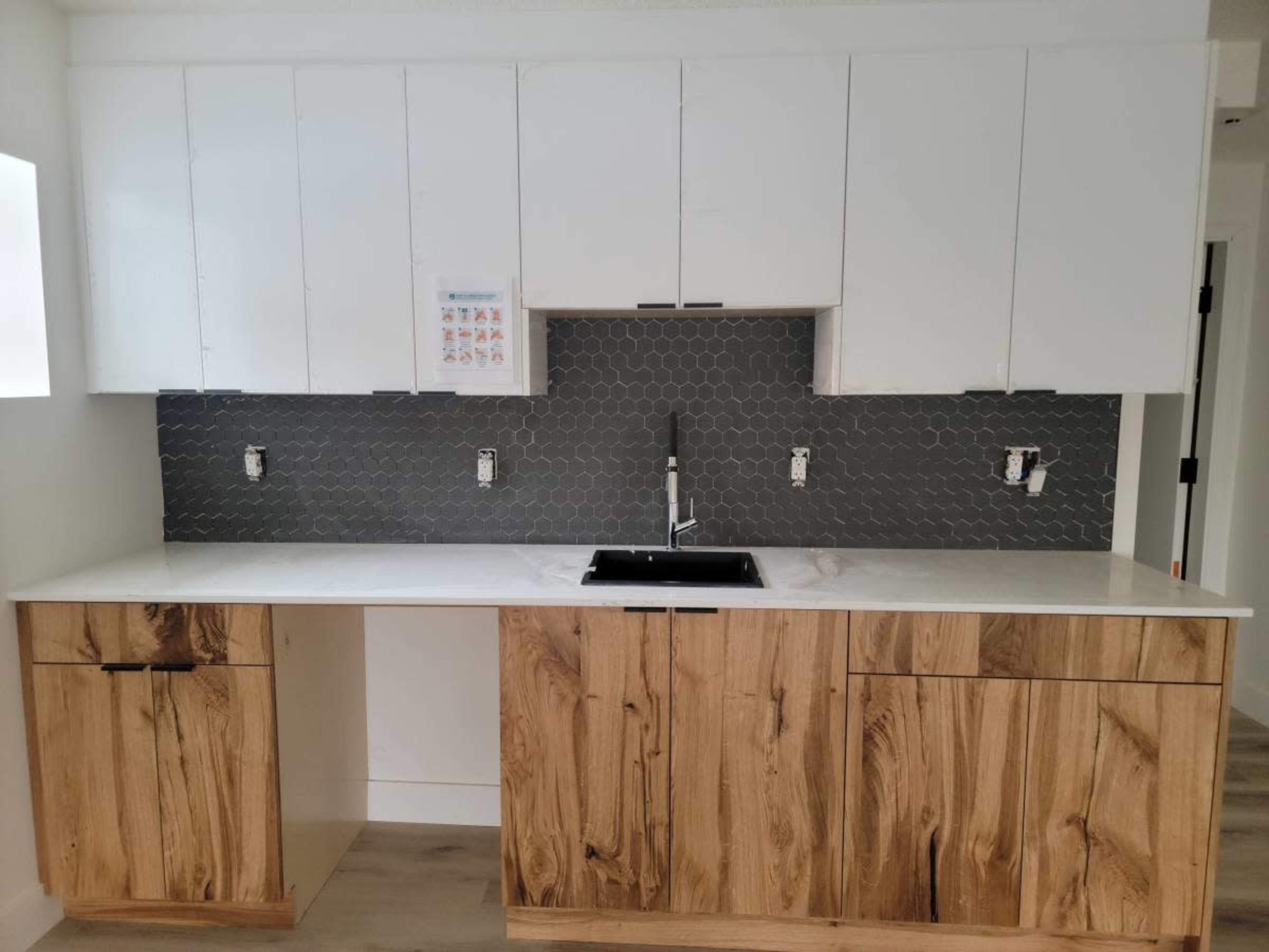 The image shows a modern kitchen with white upper cabinets, wooden lower cabinets, and a hexagonal tiled backsplash behind a black sink.