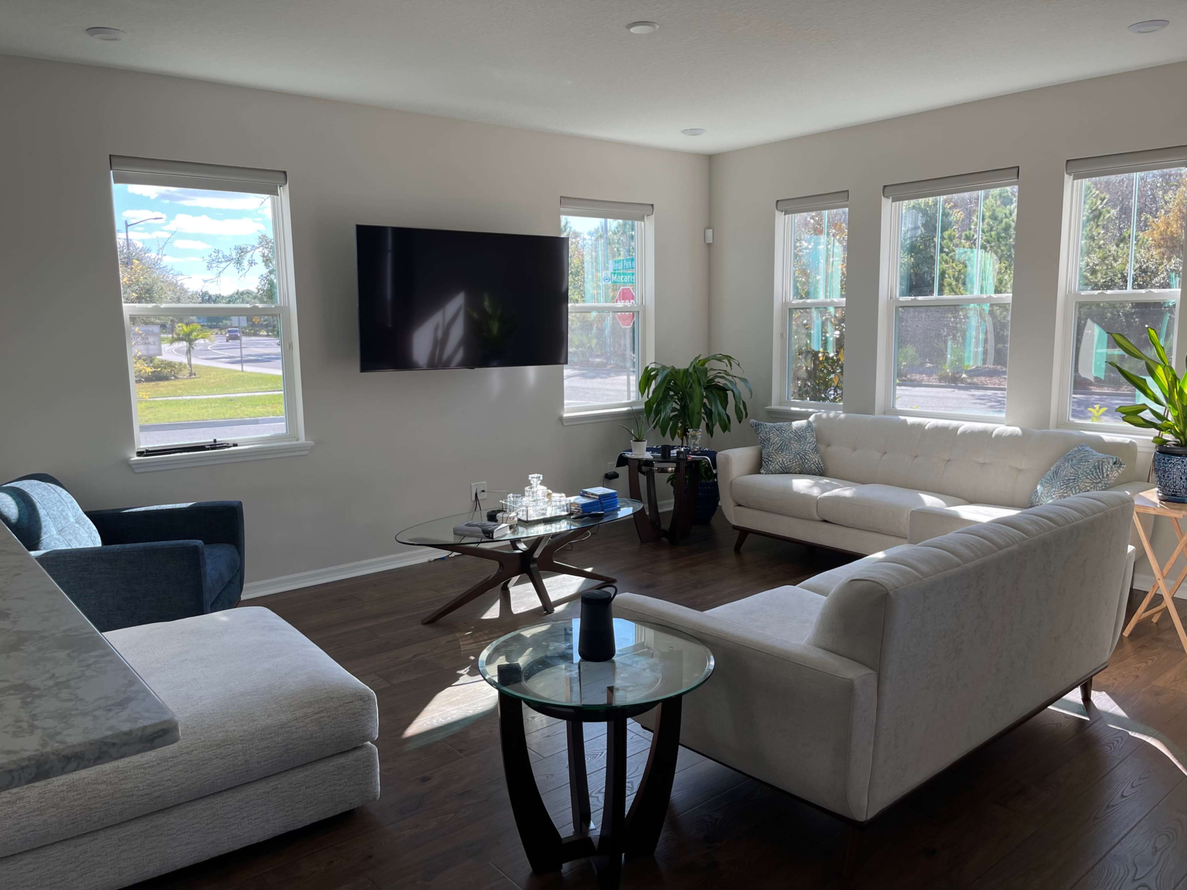 A living room features a white sofa, a blue chair, a glass coffee table, a TV mounted on the wall, and several windows allowing natural light to enter.