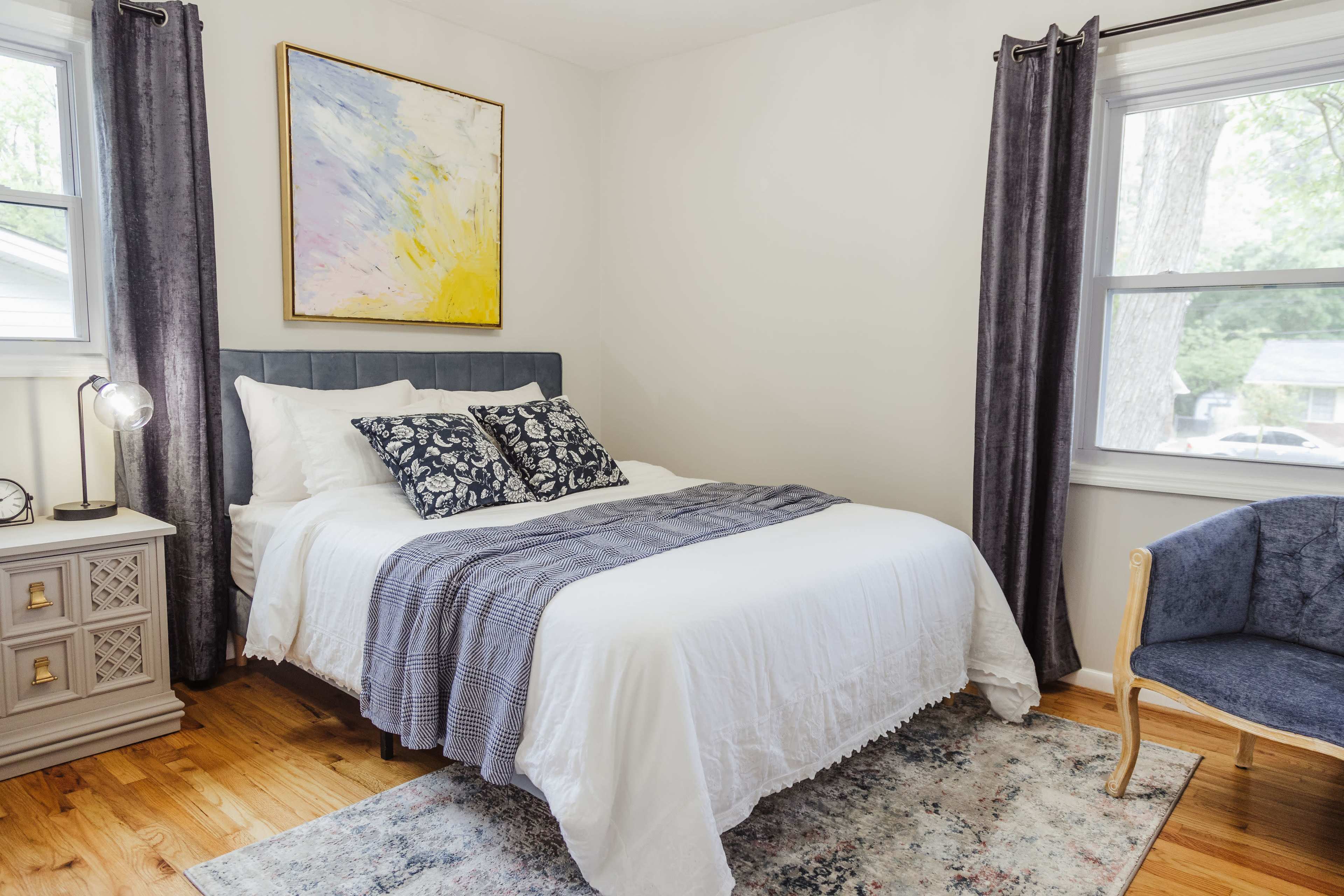 A neatly arranged bedroom features a queen-sized bed with patterned pillows, a light blanket, and a colorful piece of artwork above it, alongside a small side table and an armchair by the window.
