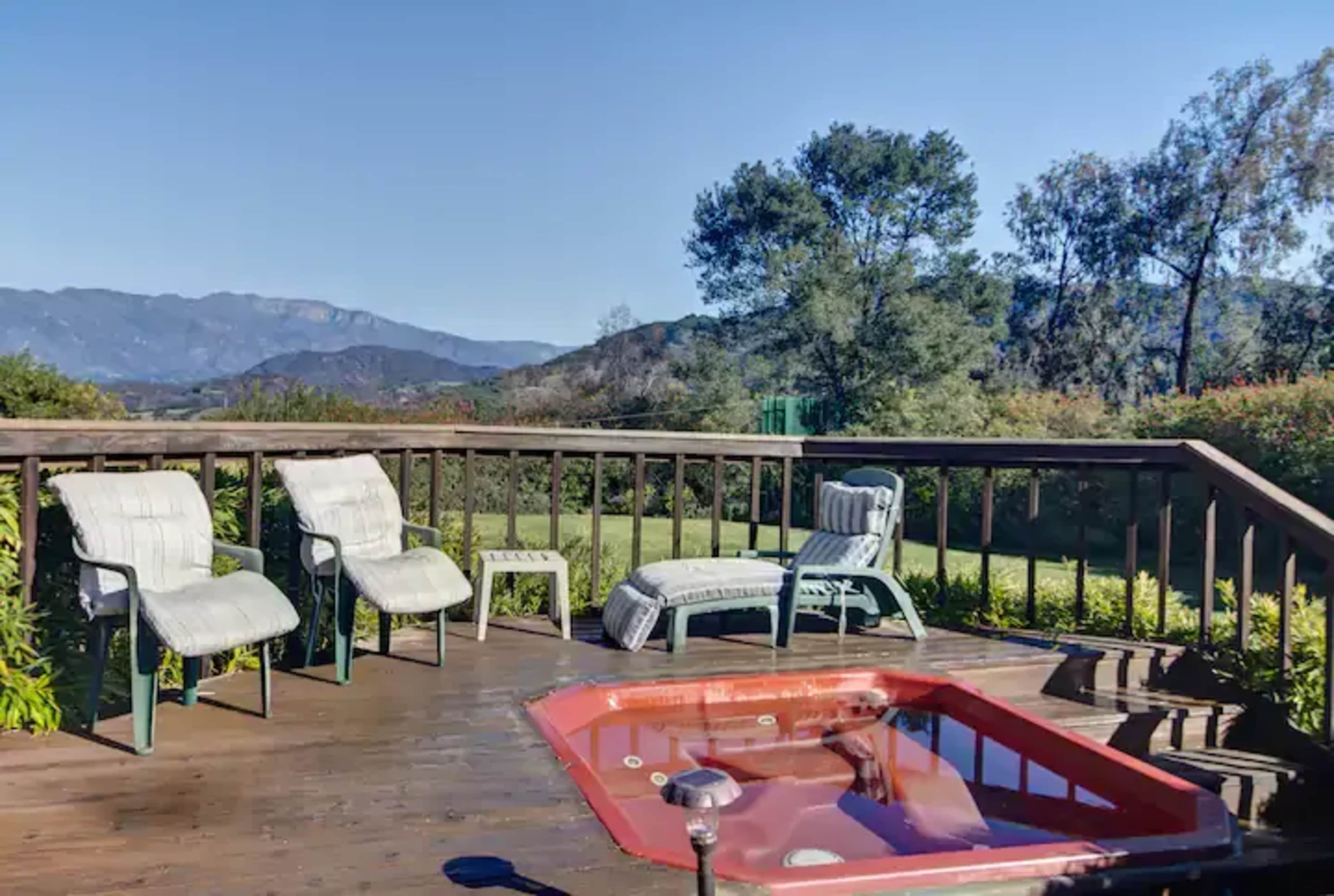 A wooden deck features a red hot tub and four chairs, set against a backdrop of mountains and clear blue sky.