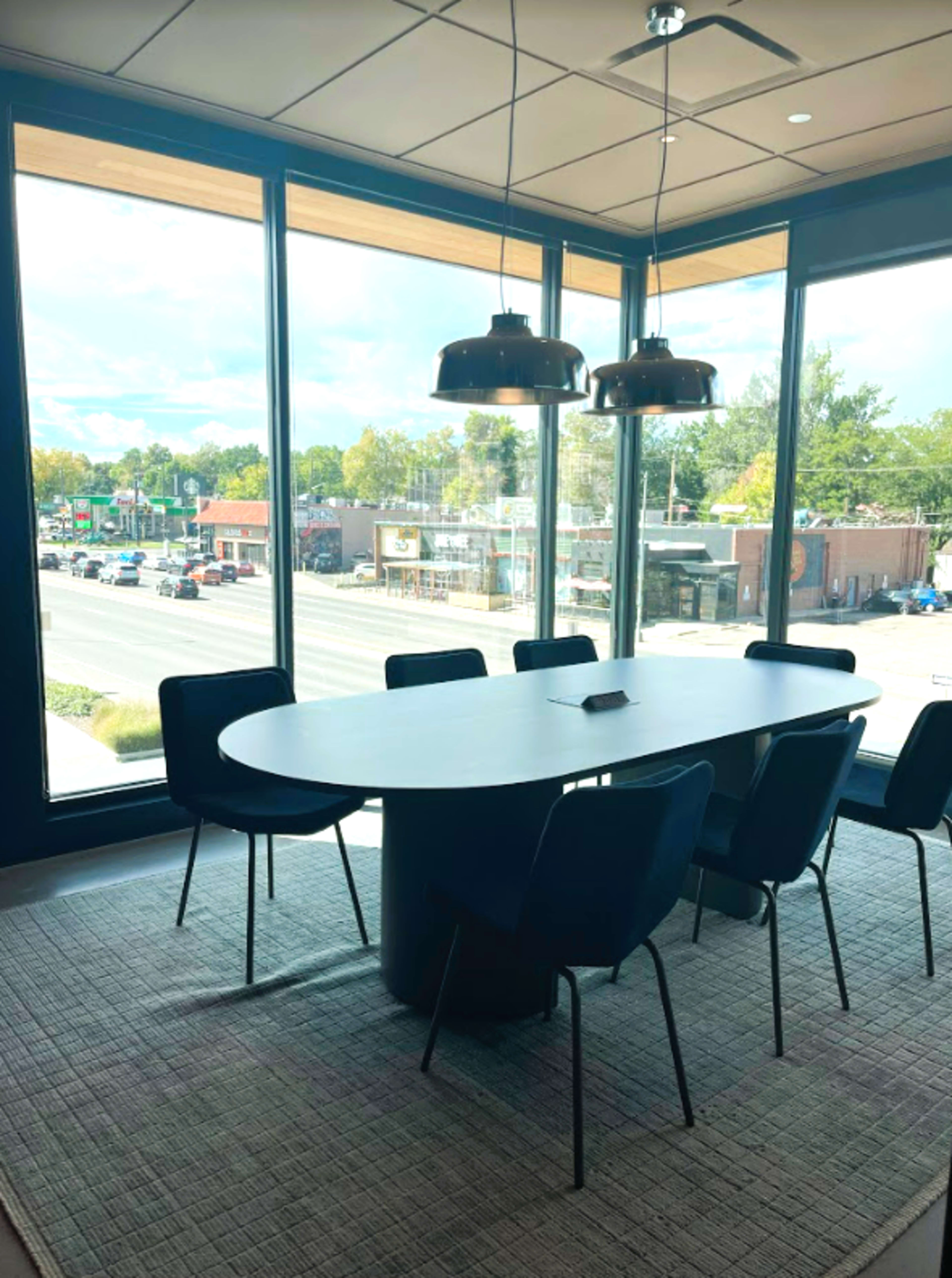 A modern conference room features a large oval table surrounded by six chairs, with large windows offering a view of a busy street and commercial buildings outside.