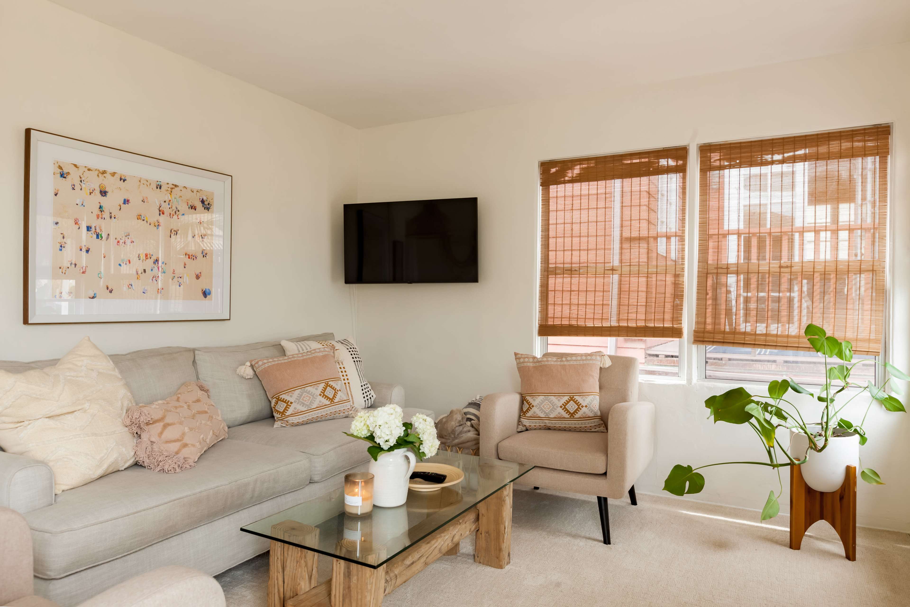 A cozy living room features a light gray sofa, two beige armchairs, a glass coffee table, a television on the wall, and large windows with bamboo shades.