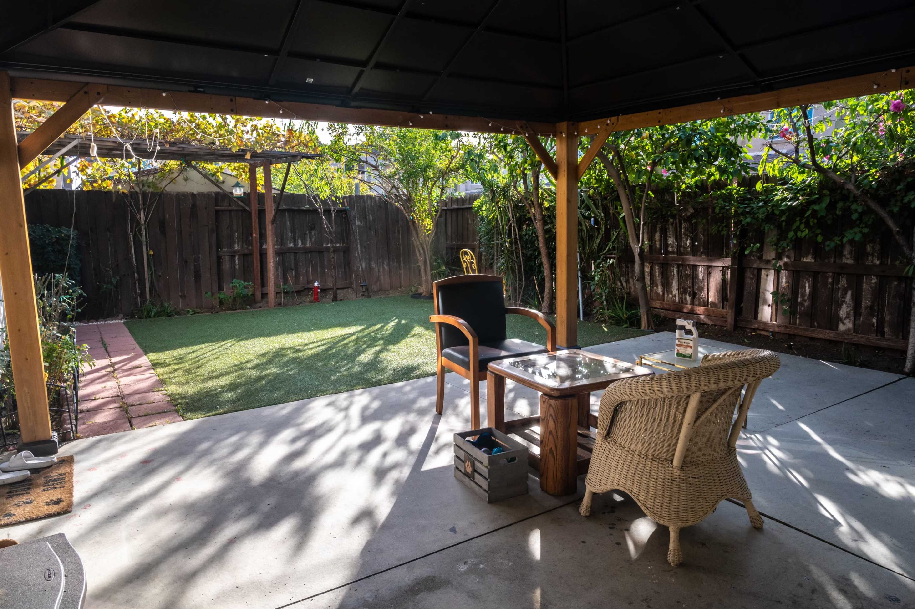 A covered patio area with a table and two chairs, surrounded by greenery and a small patch of artificial grass.