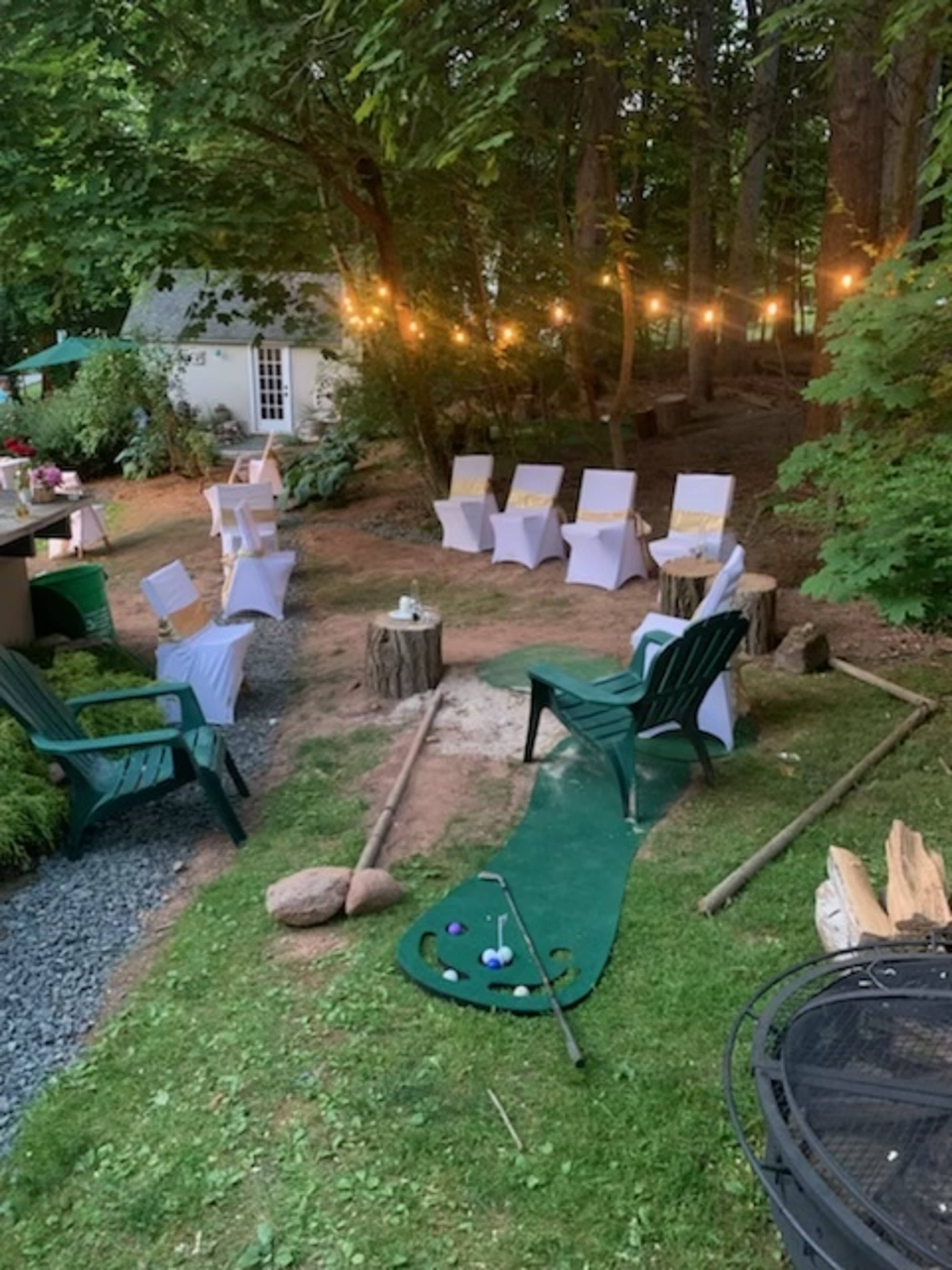 The scene shows a landscaped outdoor area with lawn chairs, string lights, a putting green, and a small table among trees.