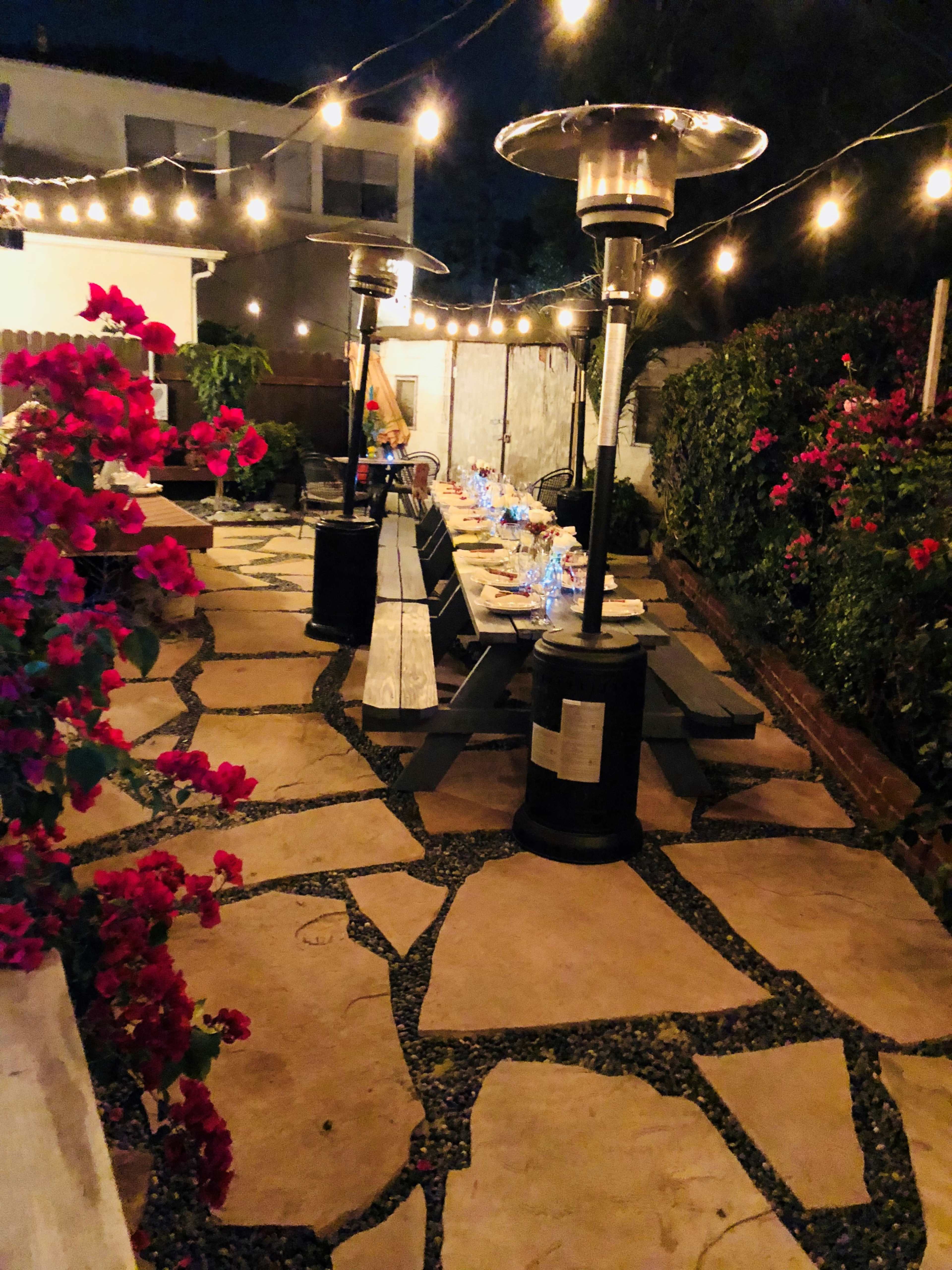 A softly lit outdoor patio area with stone pathways, patio heaters, a long dining table set with glasses, and blooming bougainvillea plants along the edges.