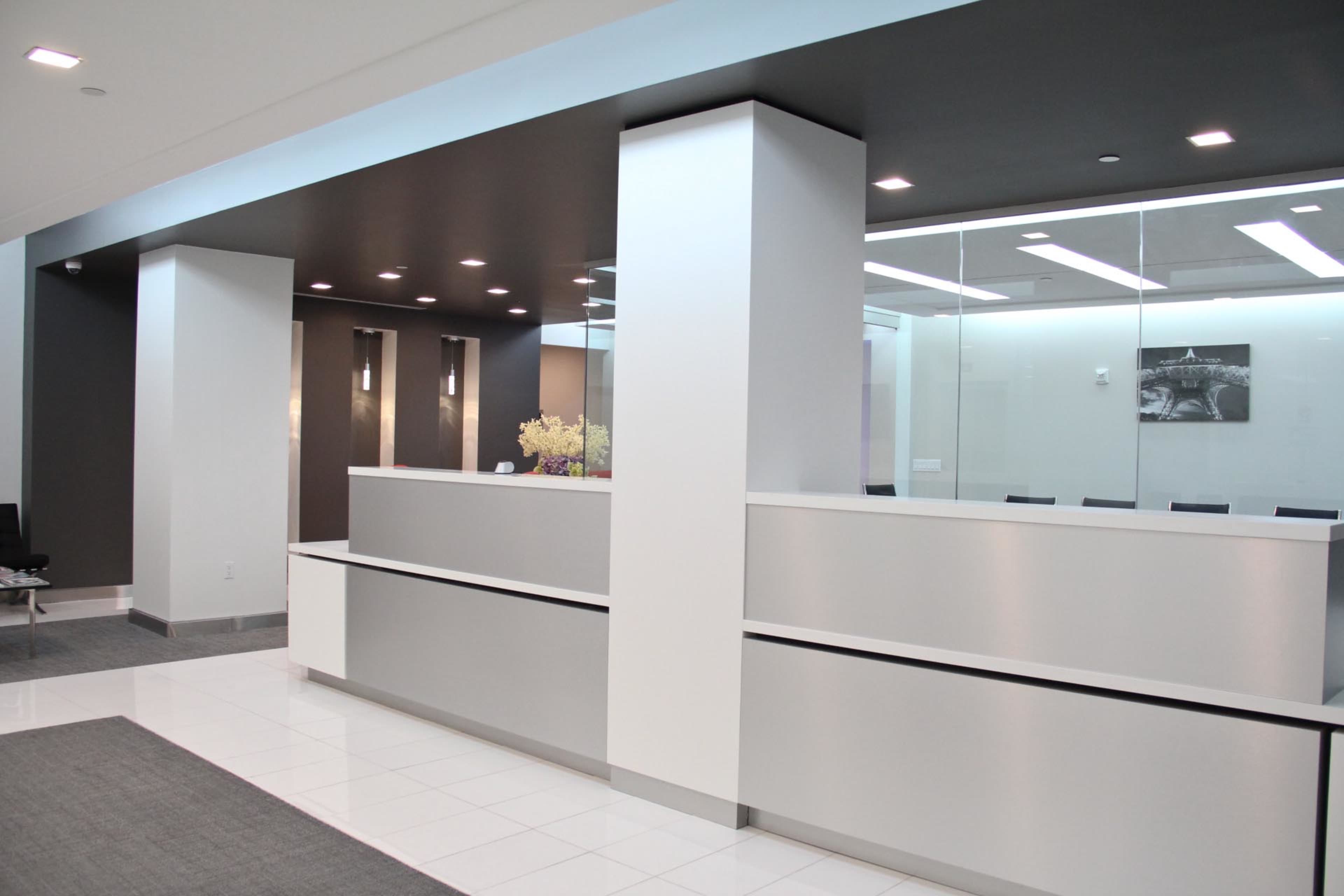 The image shows a modern reception area with sleek white and gray walls, metallic accents, and a centralized desk.