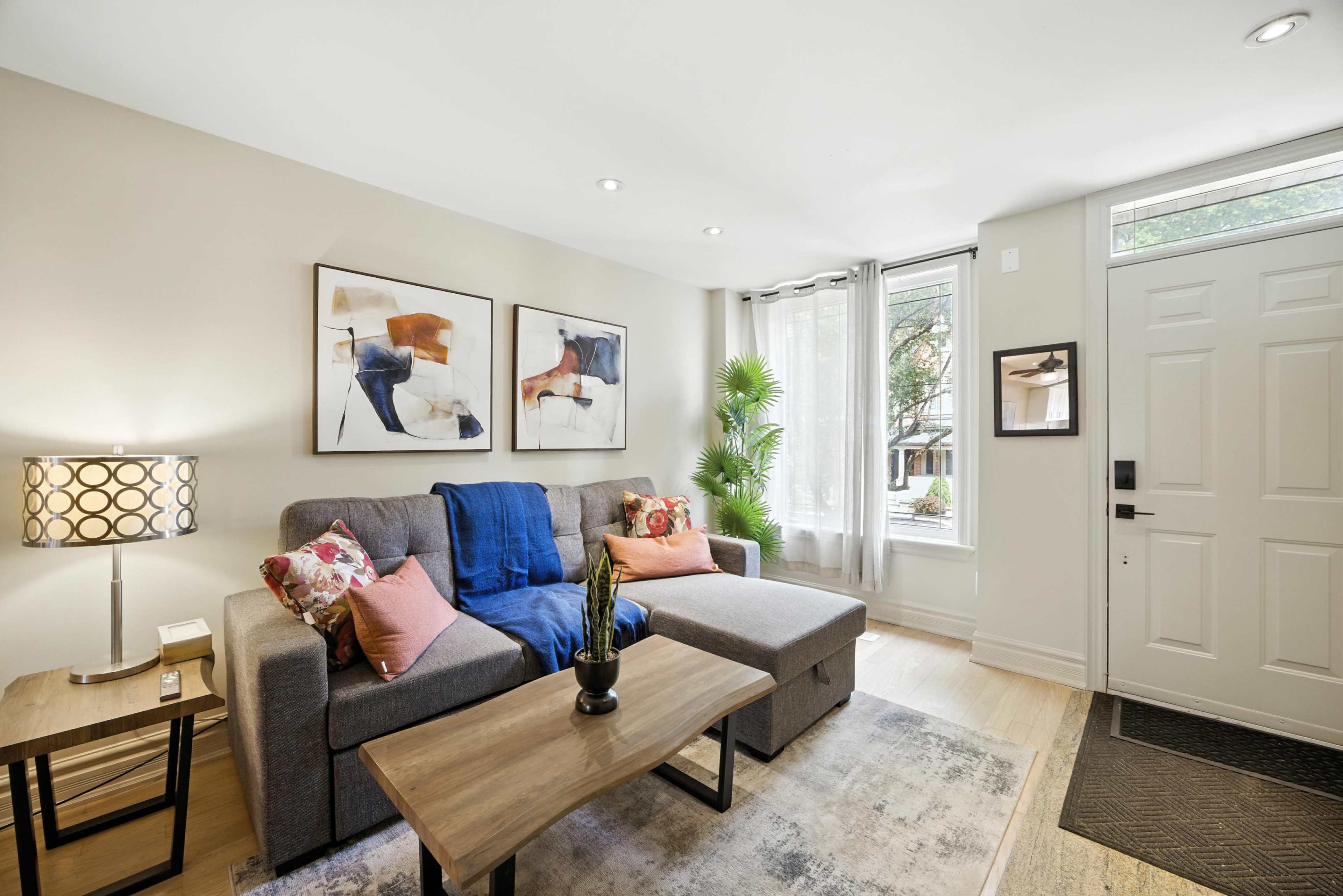 A cozy living room features a gray sectional sofa with colorful pillows, a wooden coffee table, and a welcoming entryway with a front door and windows allowing natural light.