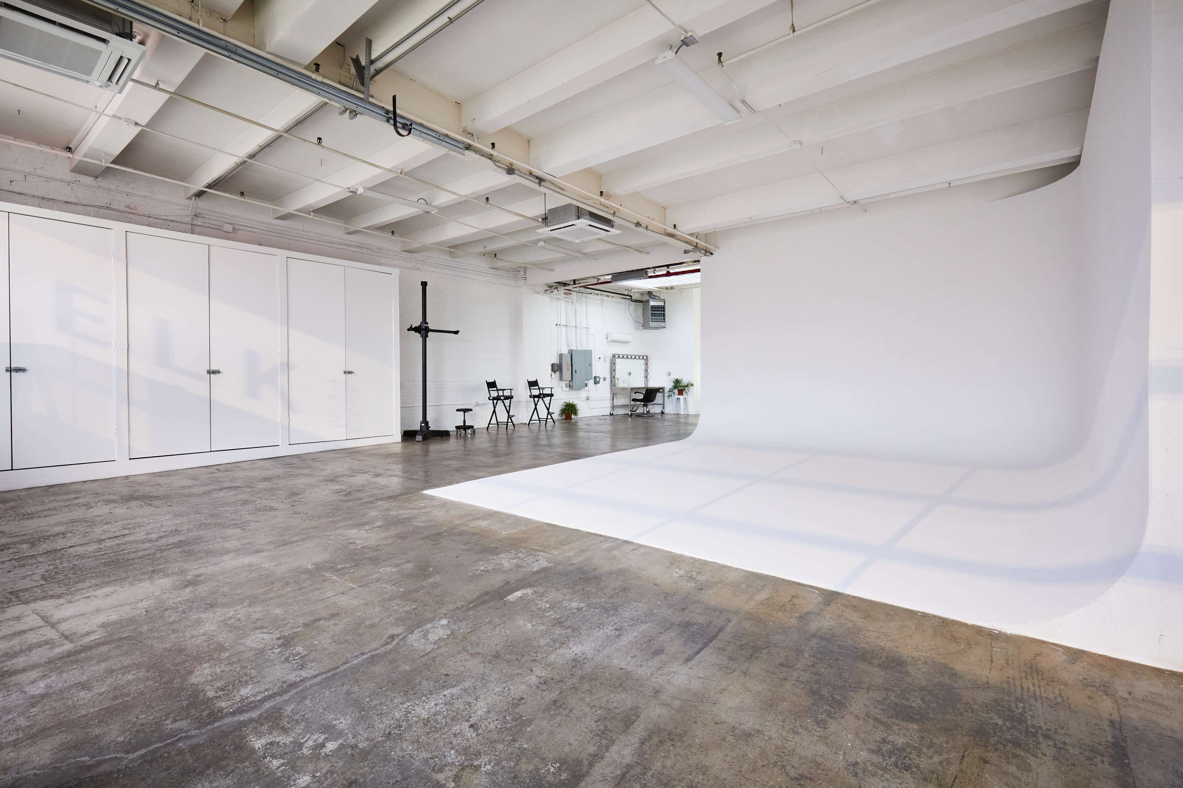 A spacious studio with a seamless white background, large windows, and concrete flooring.