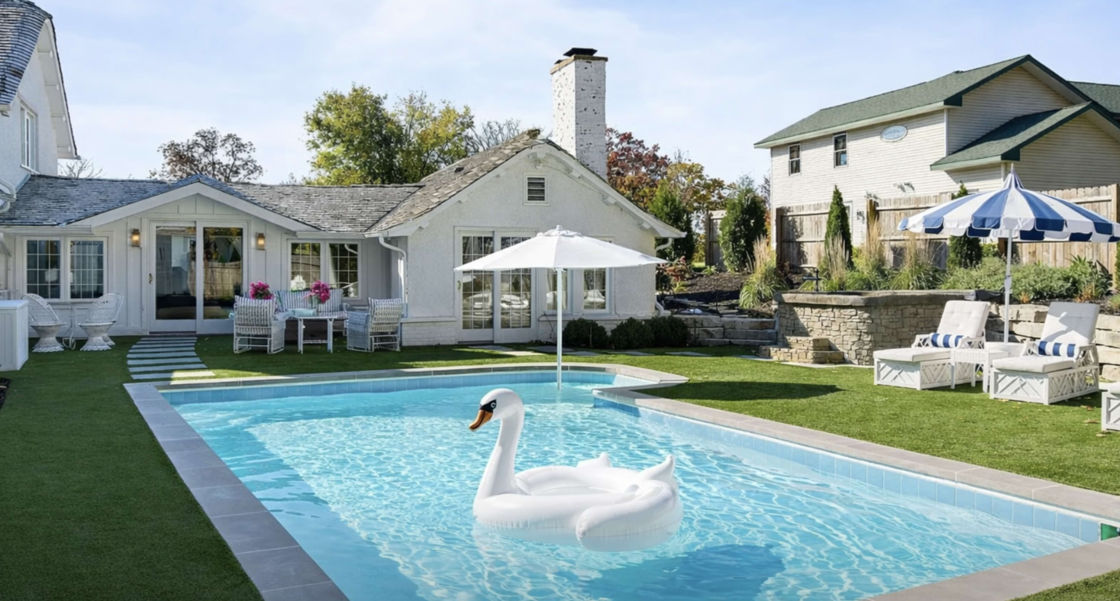 A backyard features a swimming pool with a large inflatable swan, surrounded by lounge chairs, an umbrella, and a well-maintained lawn, adjacent to a picturesque house.