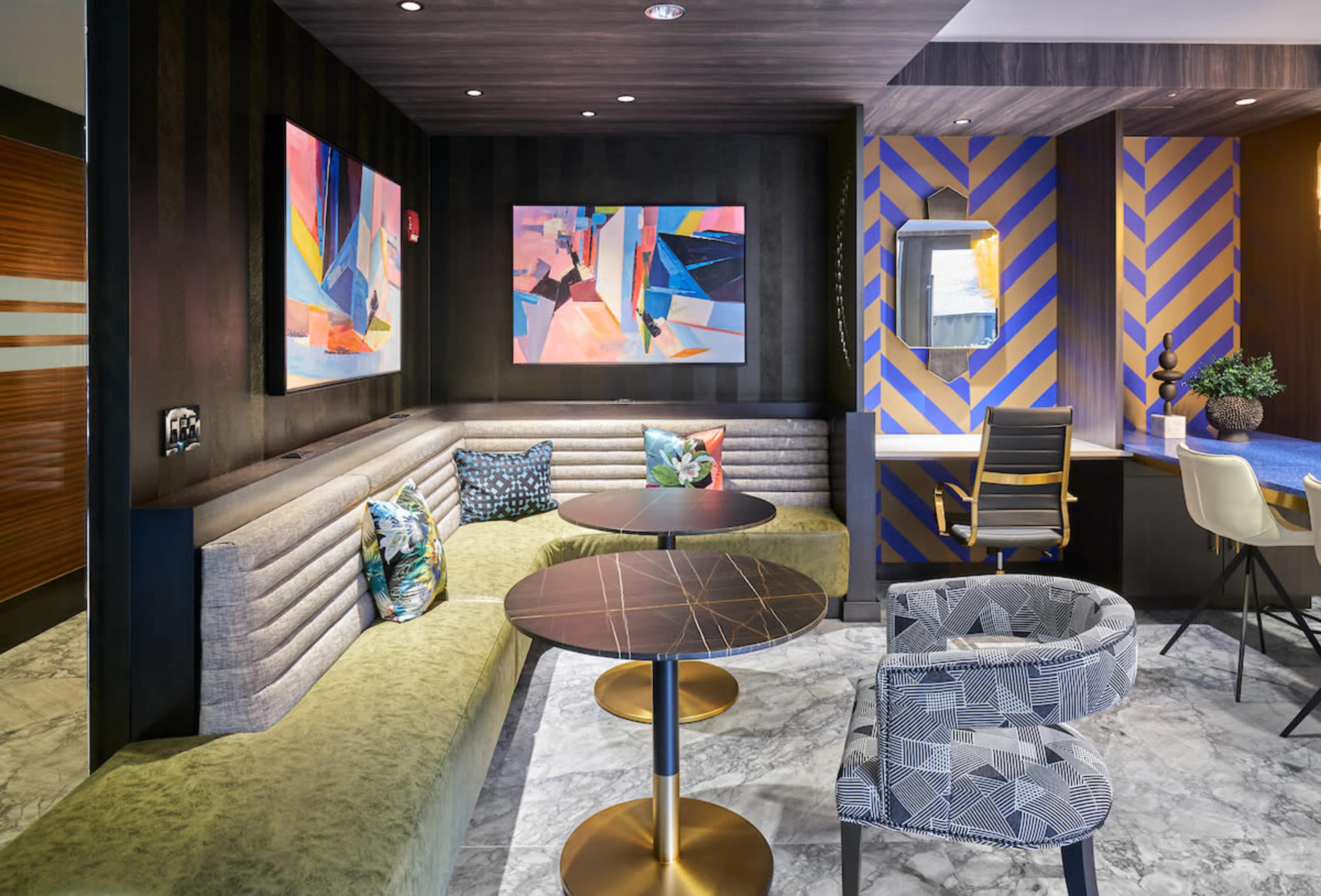 A modern lounge area features a curved green sofa, two round tables, abstract artwork on the walls, and a desk with a chair.