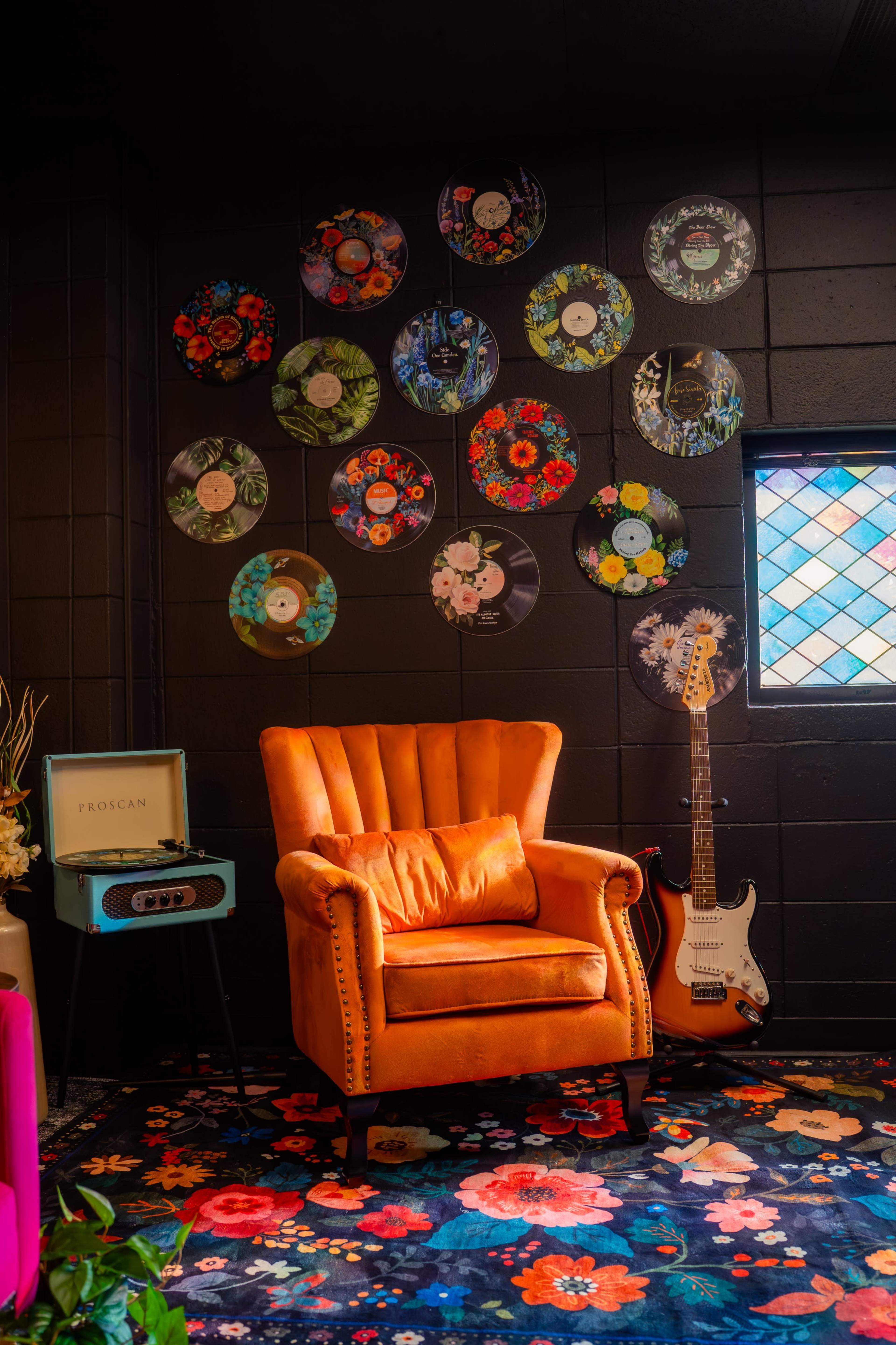 A vintage orange armchair sits against a black wall adorned with colorful vinyl records and a guitar leaning nearby.