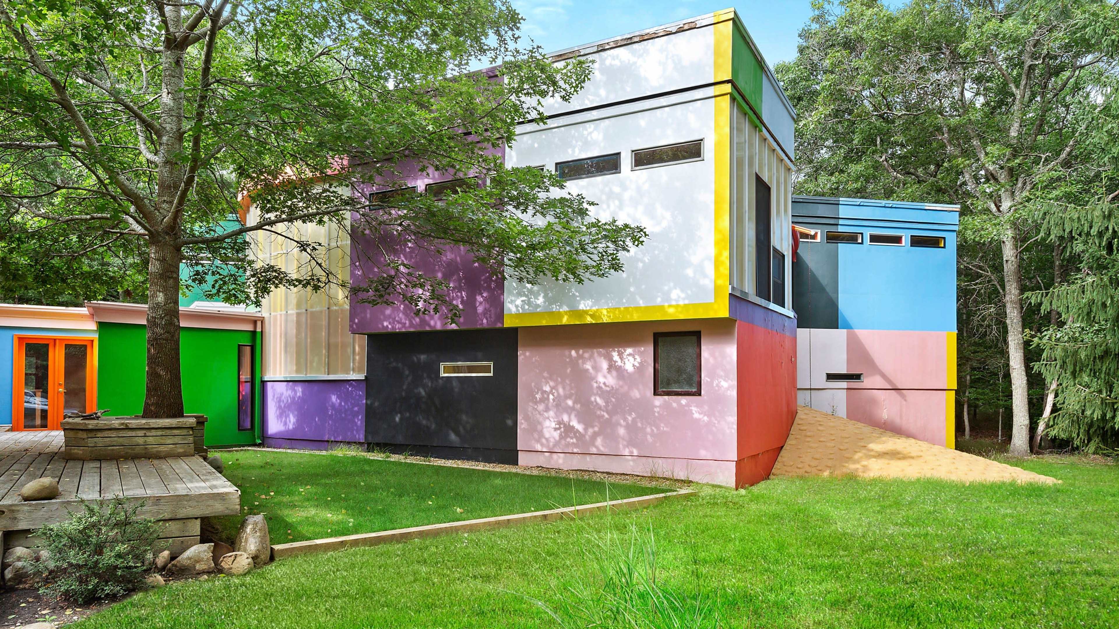 The image shows a modern house featuring geometric shapes and vibrant colors, surrounded by green grass and trees.