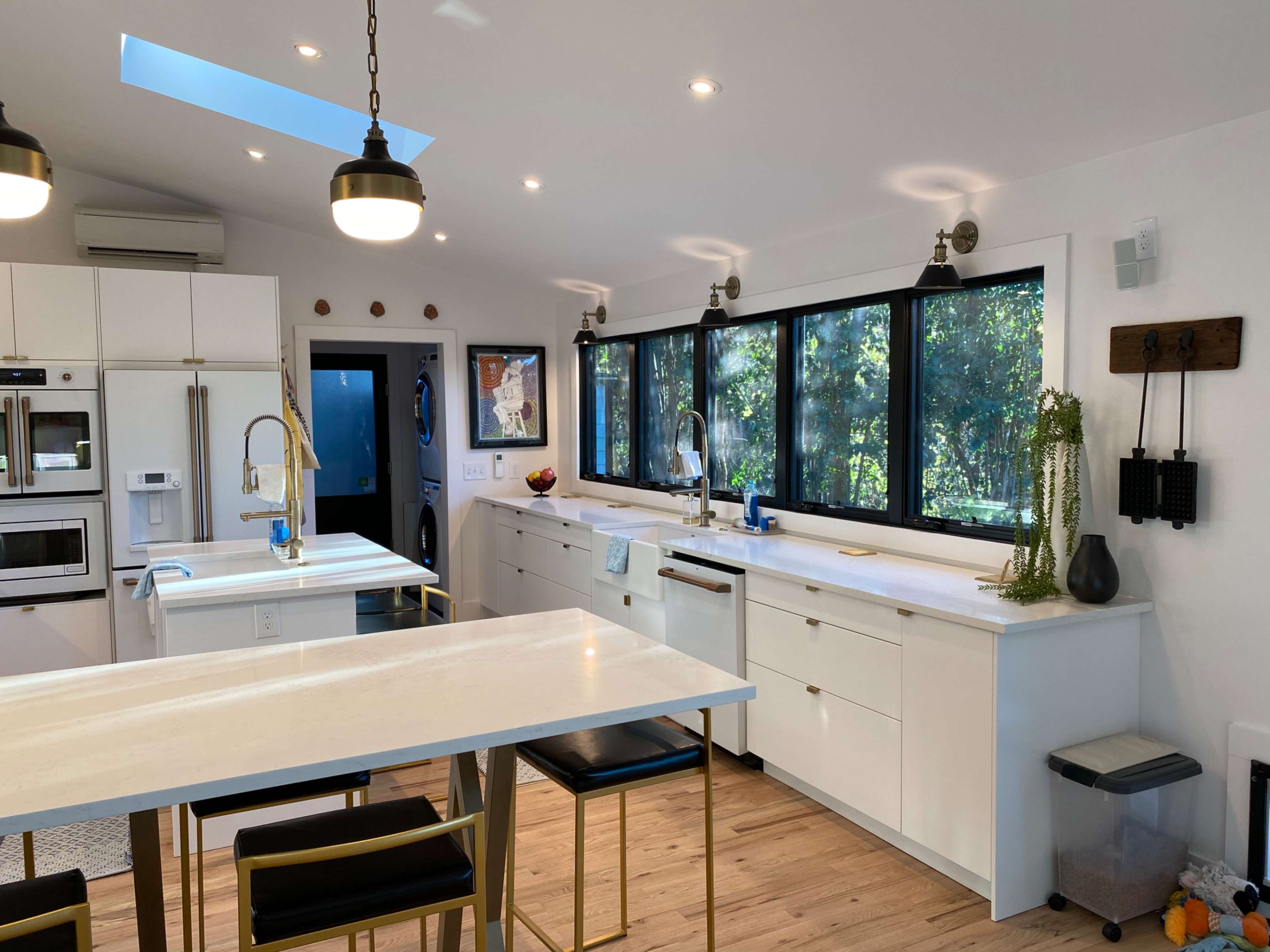 The image shows a modern kitchen with a white color scheme, large windows, and a central island featuring bar stools.