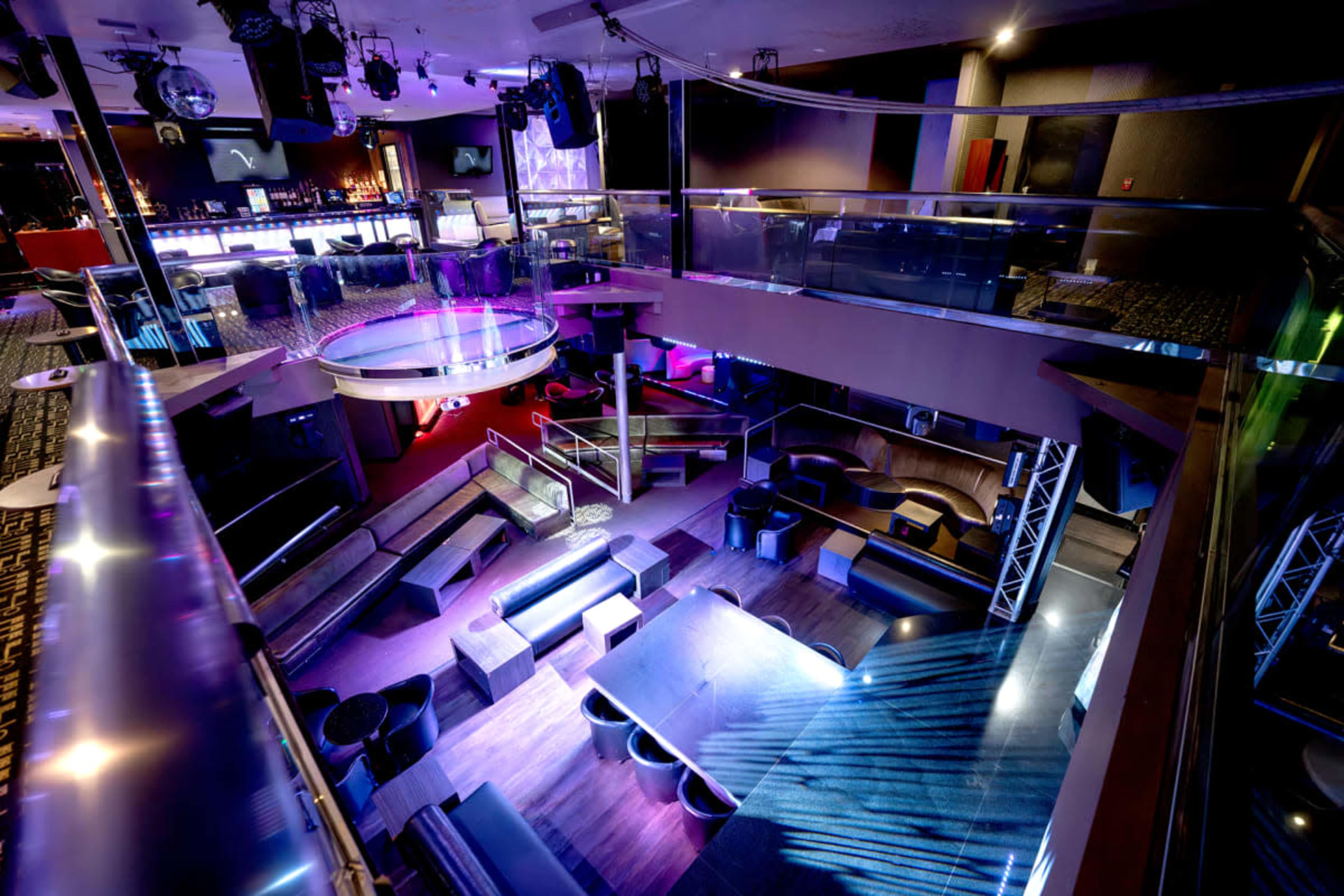 The image shows a modern nightclub interior with multiple seating areas, a central dance floor, and colorful lighting.