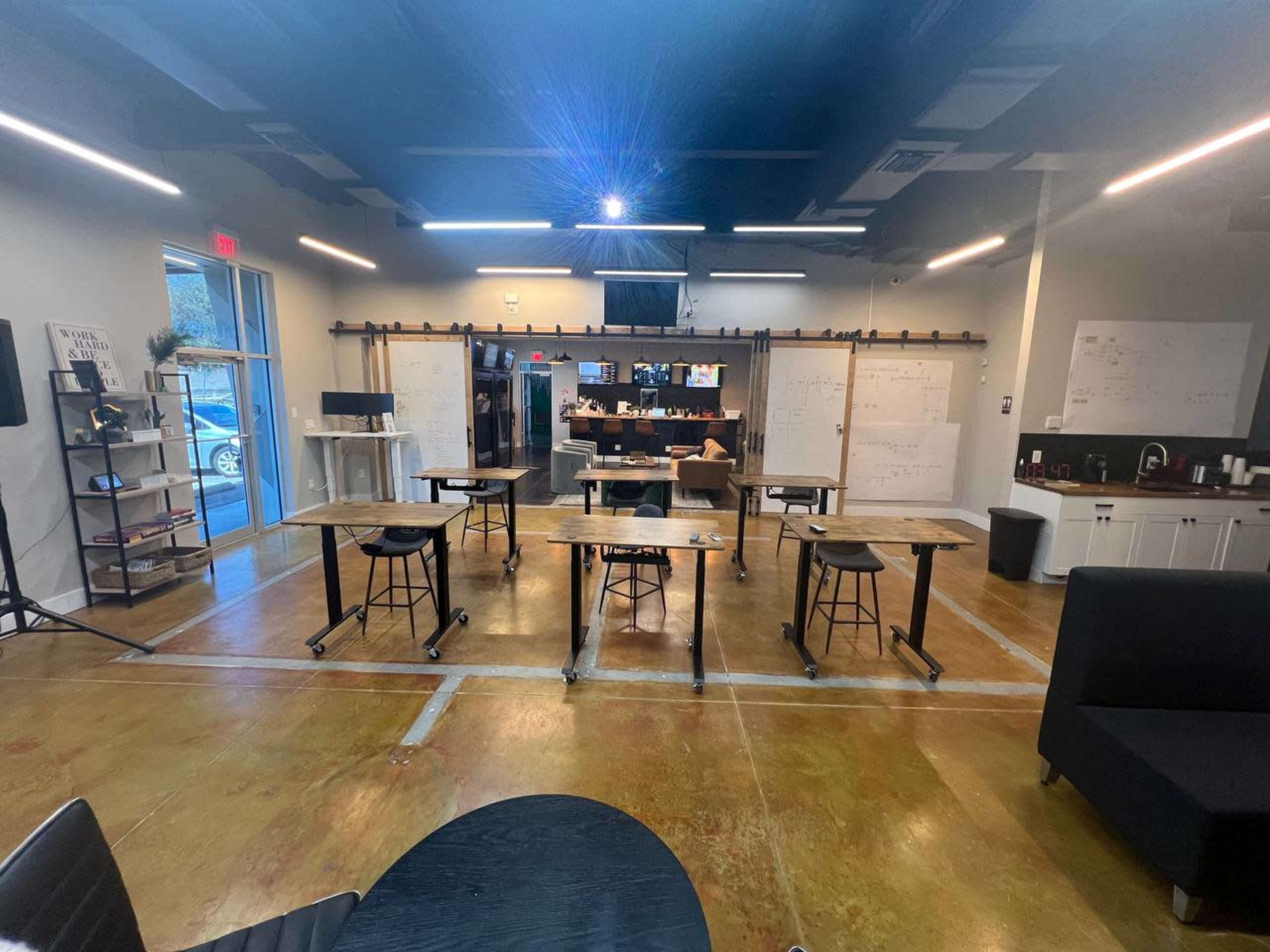 A modern workspace with several tables and chairs arranged, whiteboards mounted on the walls, and a kitchenette area in the background.