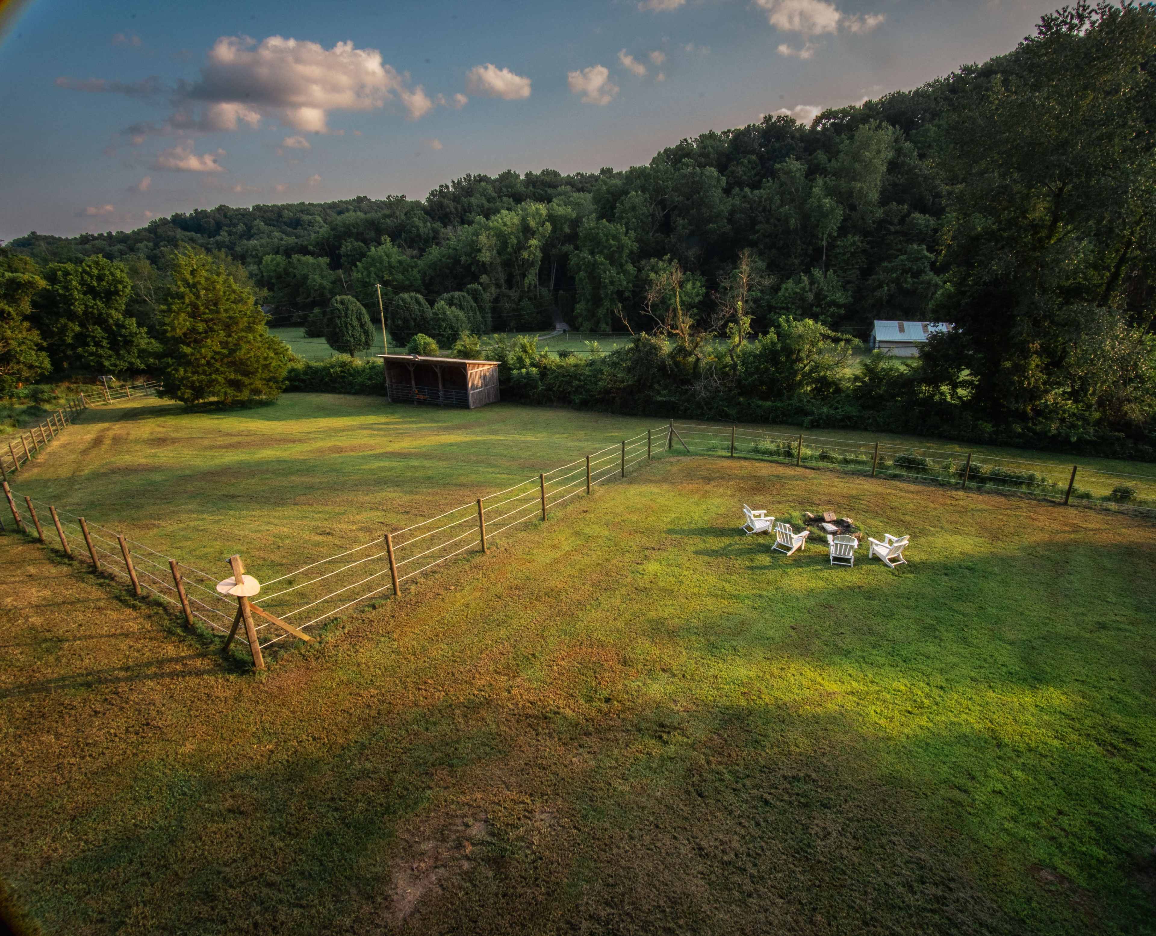 The image shows a grassy field surrounded by trees, with a seating area featuring white chairs and a fire pit near a wooden structure in the background.