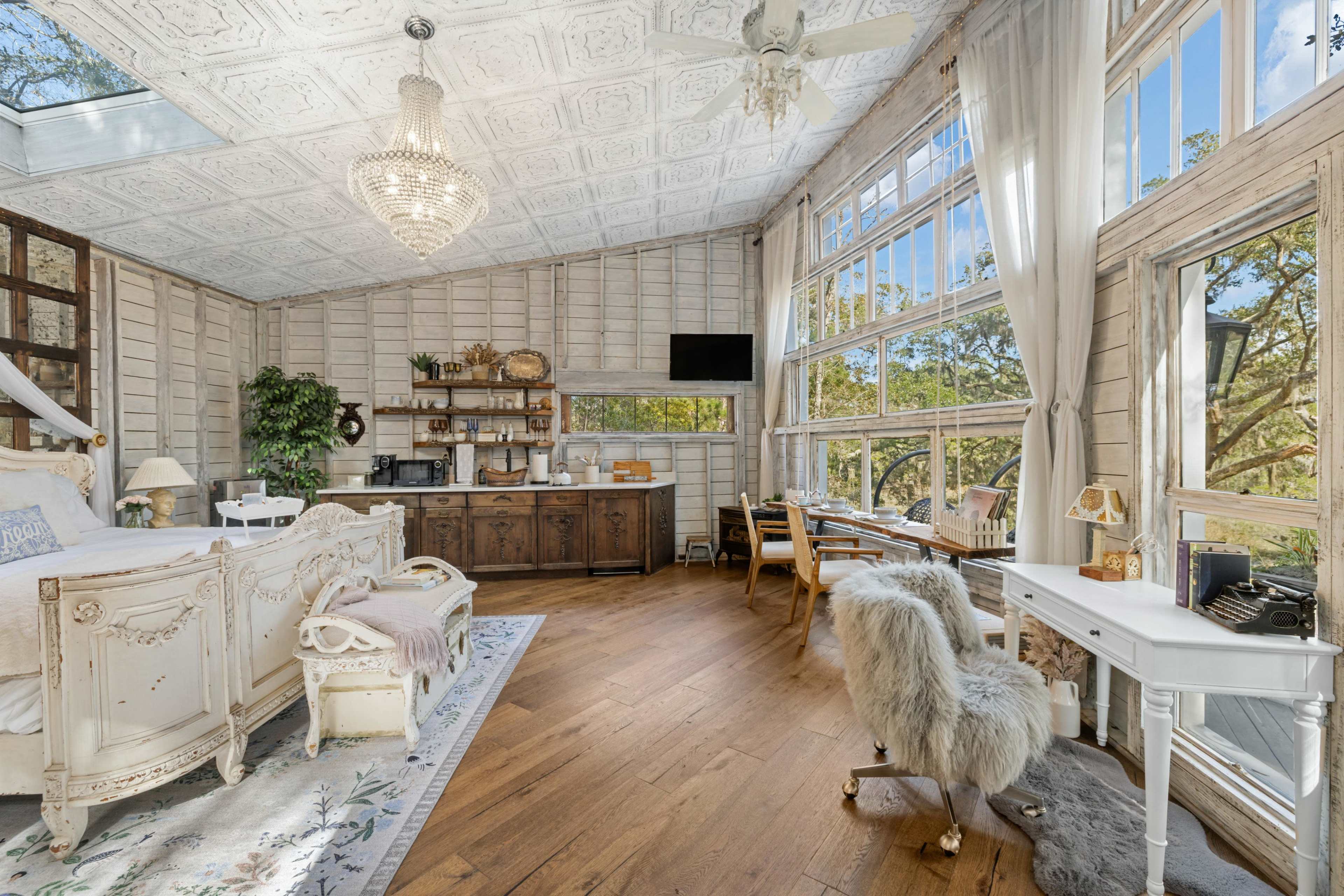 A bright, airy room with large windows, wooden flooring, and a mix of vintage furniture, including a seating area, a small kitchen space, and a workspace.