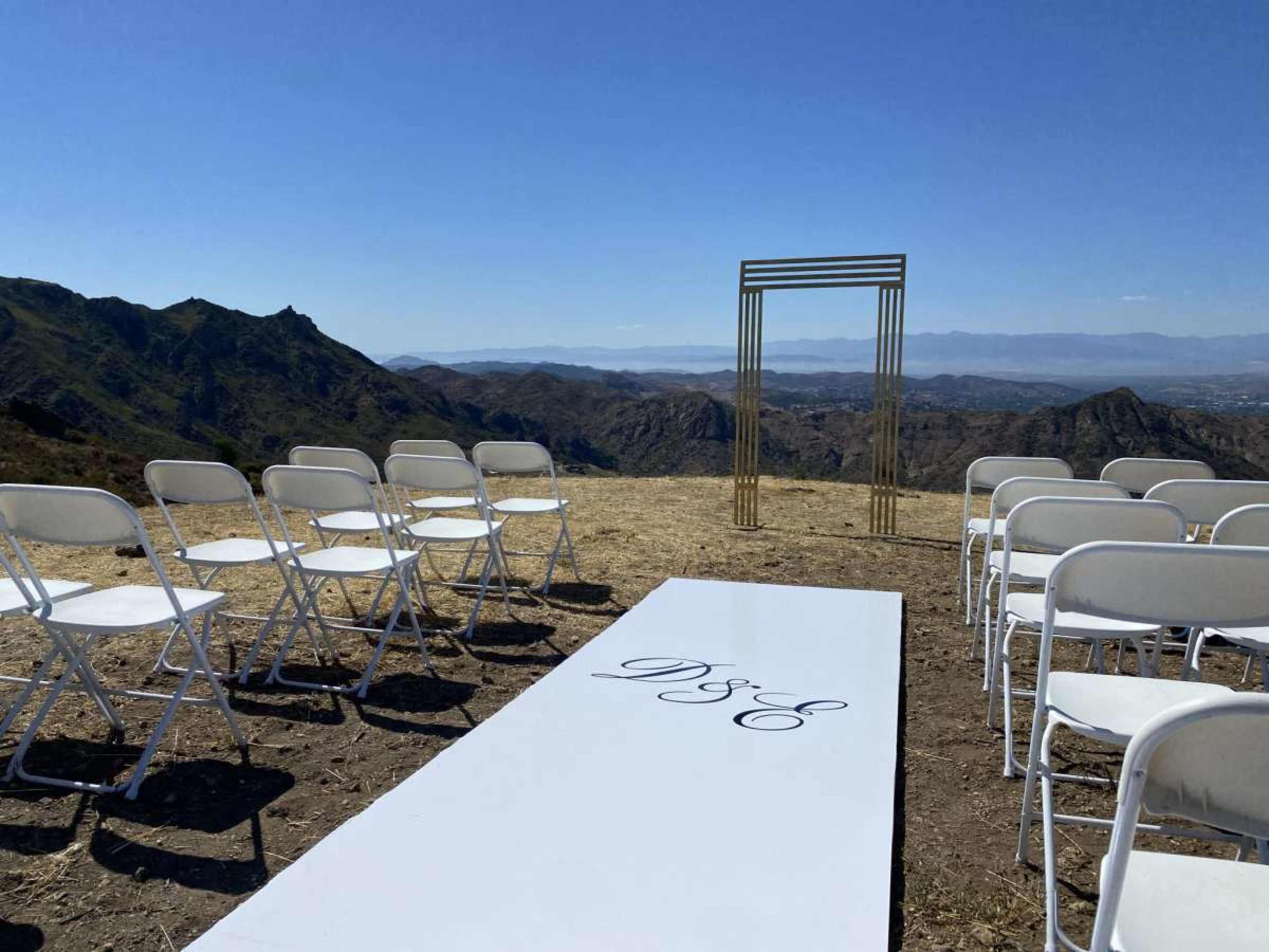 A wedding ceremony setup features white chairs arranged on a mountain peak, with a pathway leading to a golden archway against a backdrop of rolling hills.