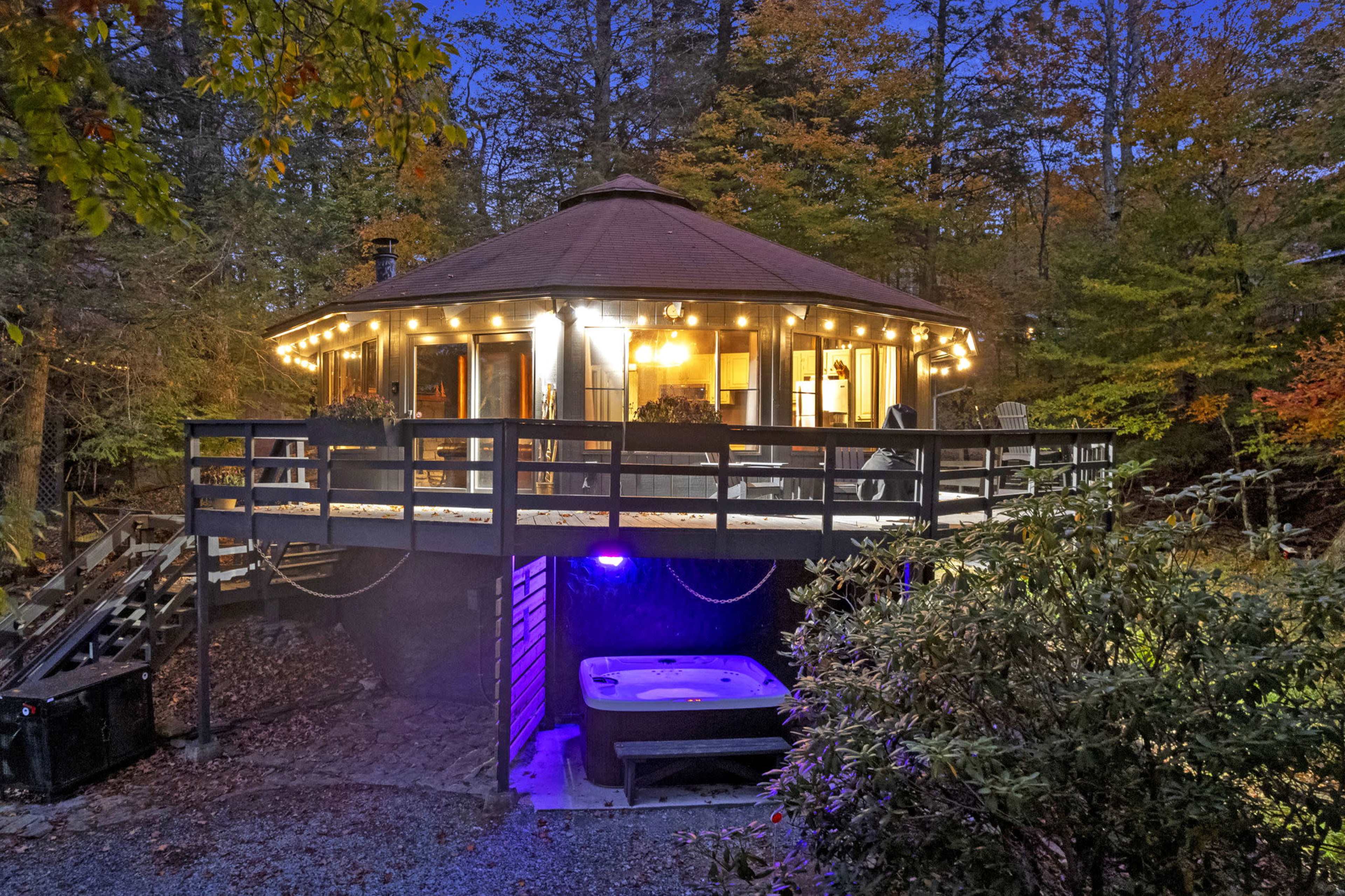 A circular, wooden cabin with a deck is illuminated by string lights and features a hot tub underneath at dusk, surrounded by trees.