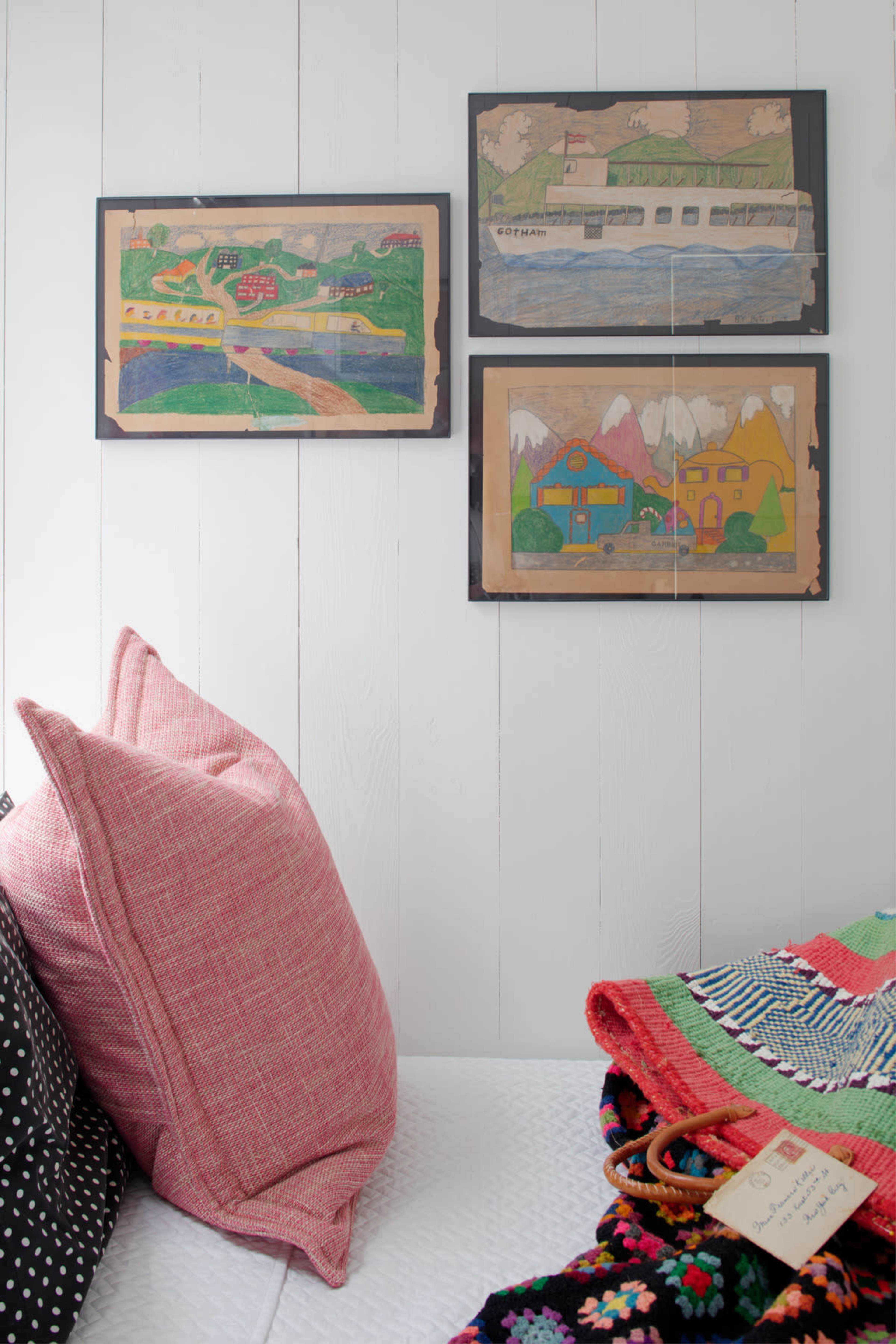 The image shows a cozy corner of a room featuring a white wall adorned with three framed colorful illustrations, accompanied by a bed with patterned pillows and a vibrant textile blanket.