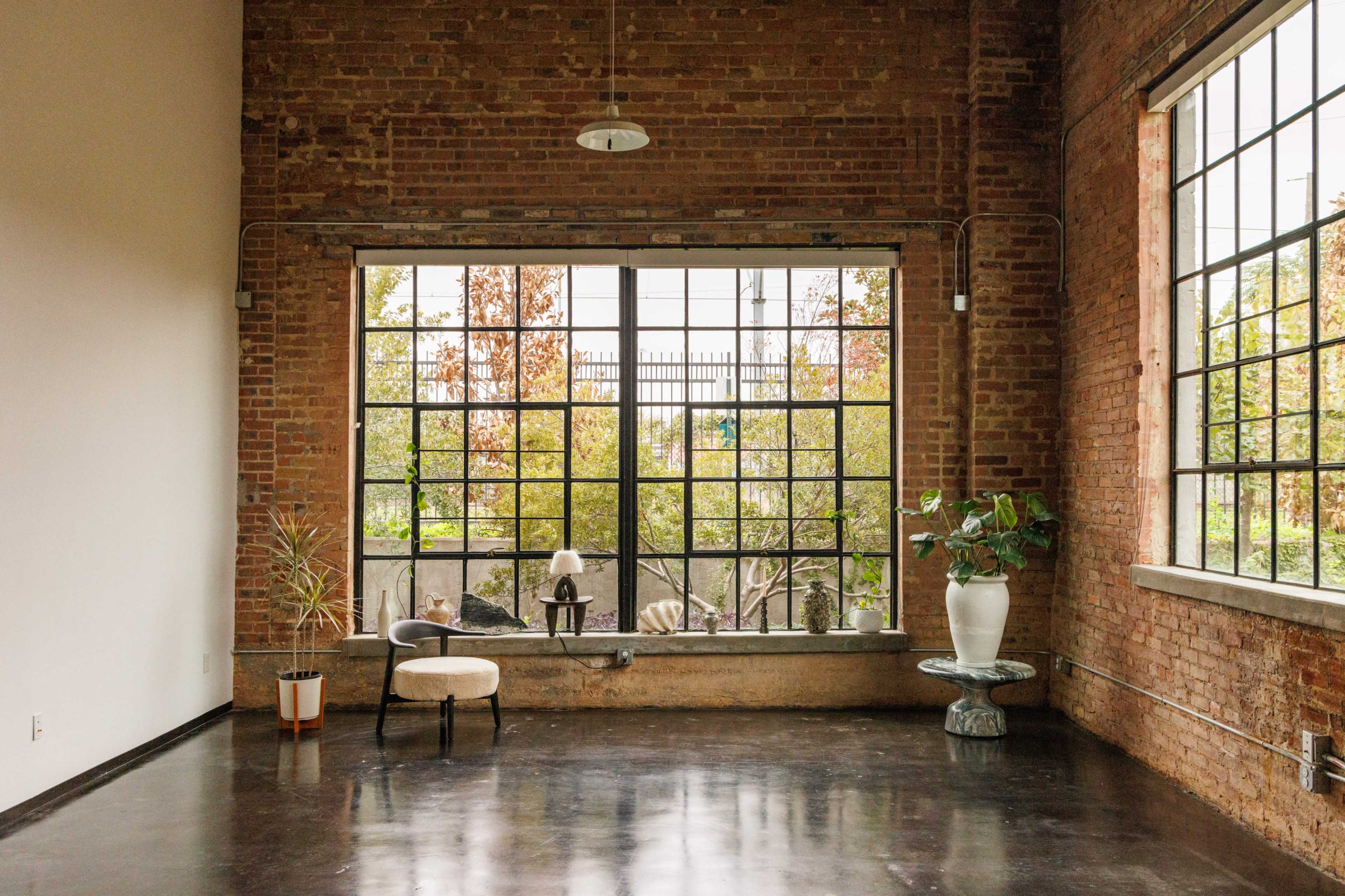 The image shows a spacious, industrial-style room with exposed brick walls and large windows allowing natural light to enter, featuring minimal furnishings.