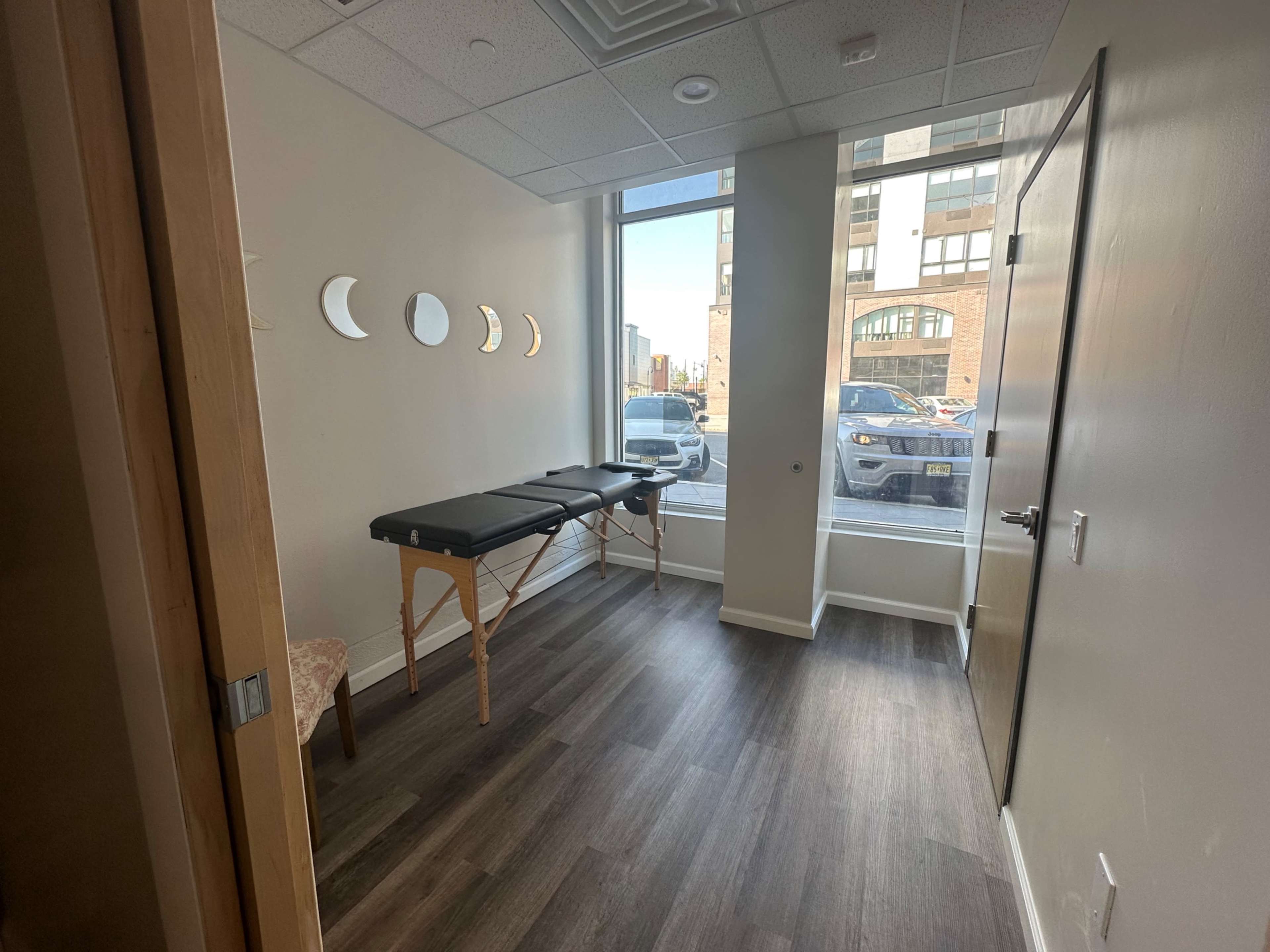 A treatment room with a massage table, a chair, and large windows facing an exterior view of cars and buildings.