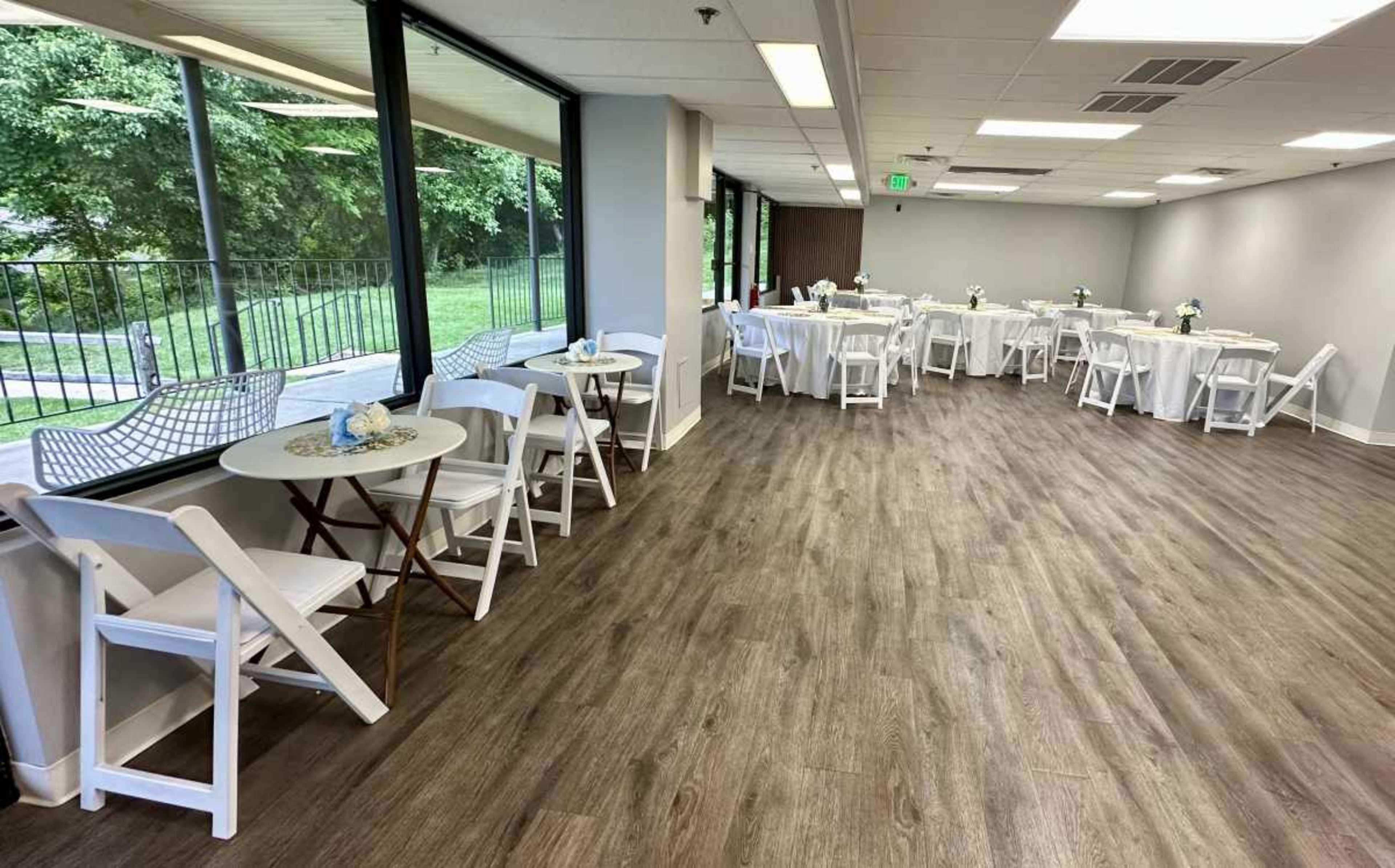 The image shows a spacious event room with white tables and chairs arranged neatly, large windows overlooking greenery, and a well-lit interior.
