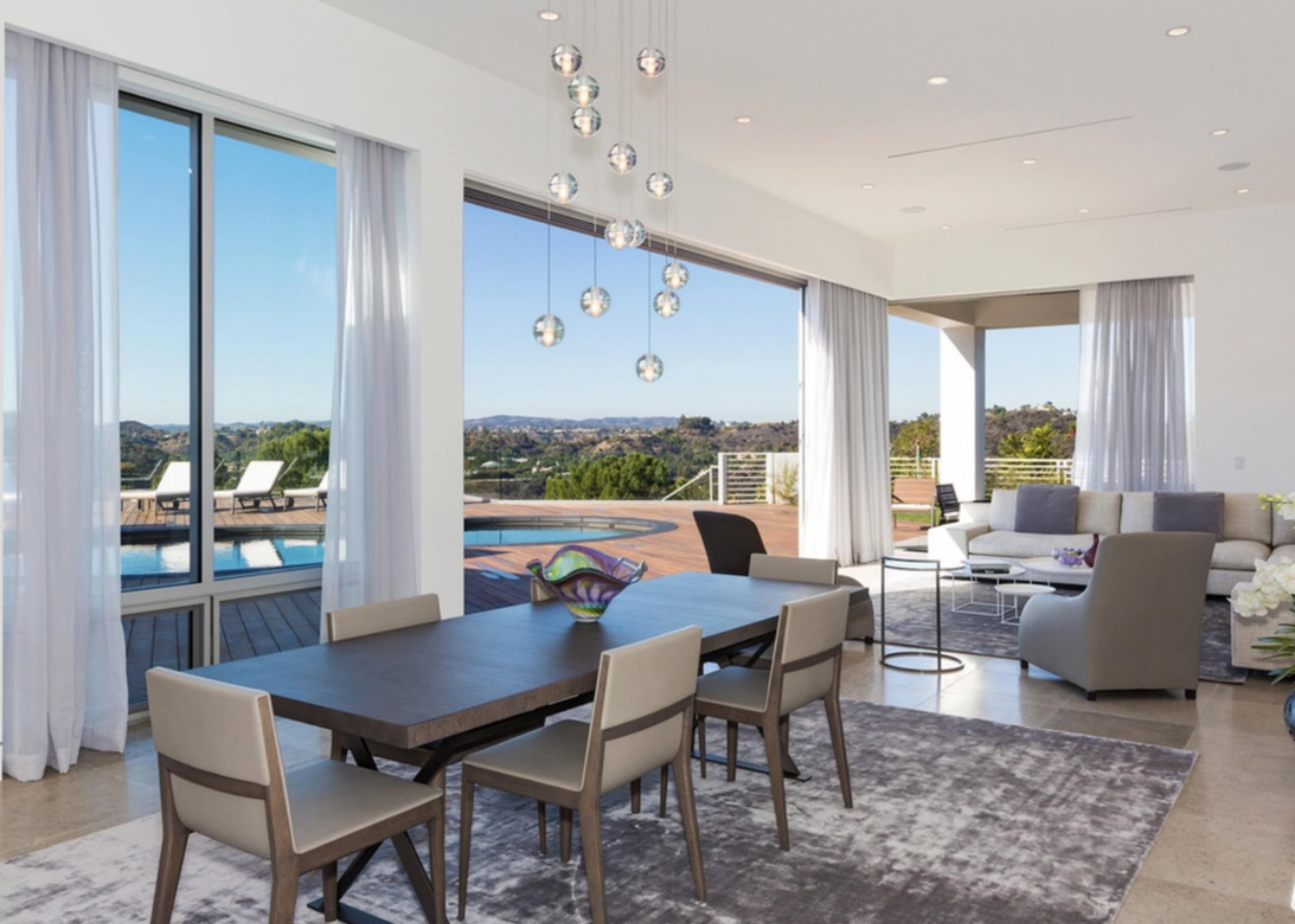 A modern dining area features a large table surrounded by chairs, with expansive windows overlooking a pool and scenic view.