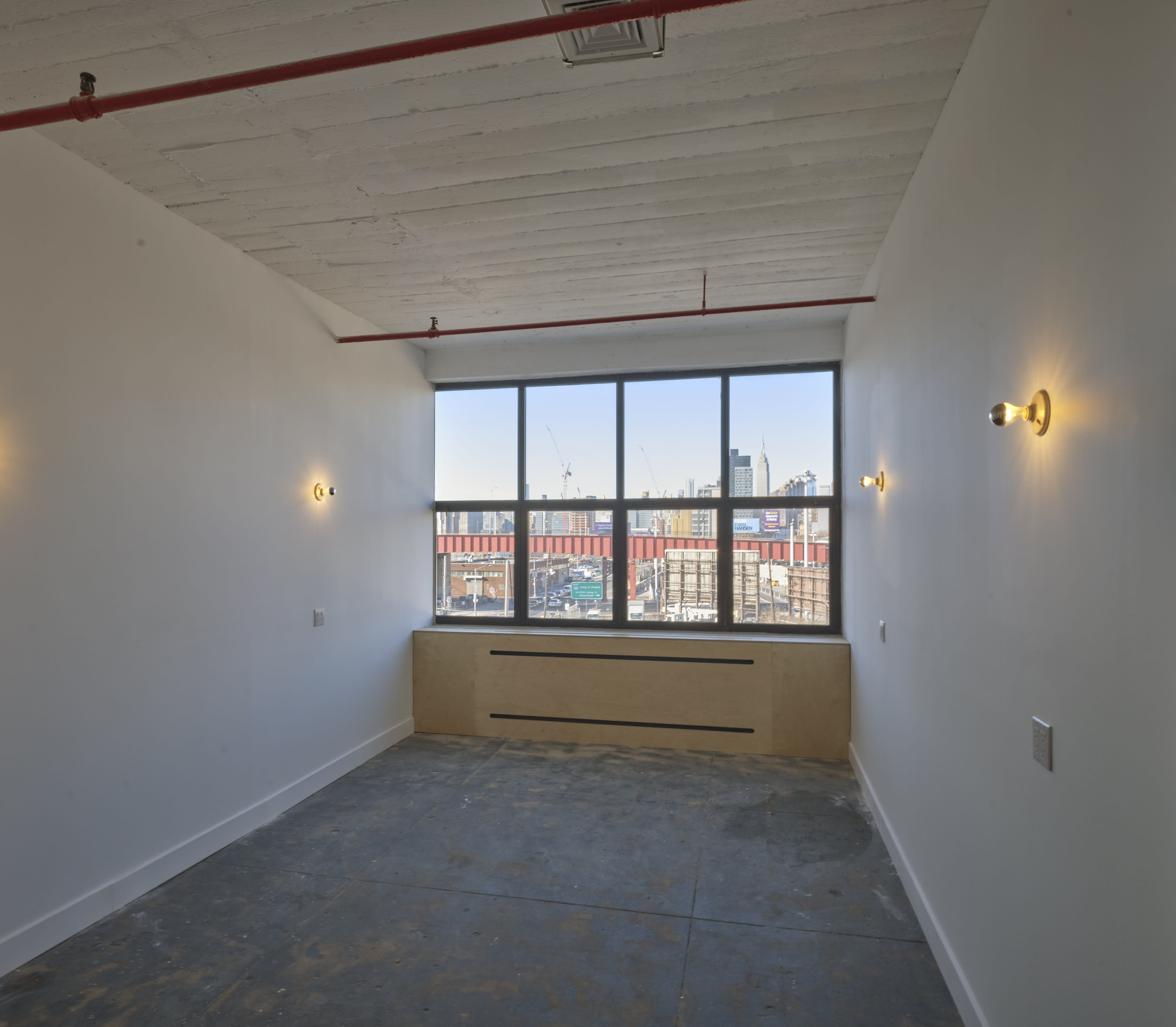 An empty room with a large window overlooking a cityscape, featuring exposed red piping and unfinished concrete flooring.