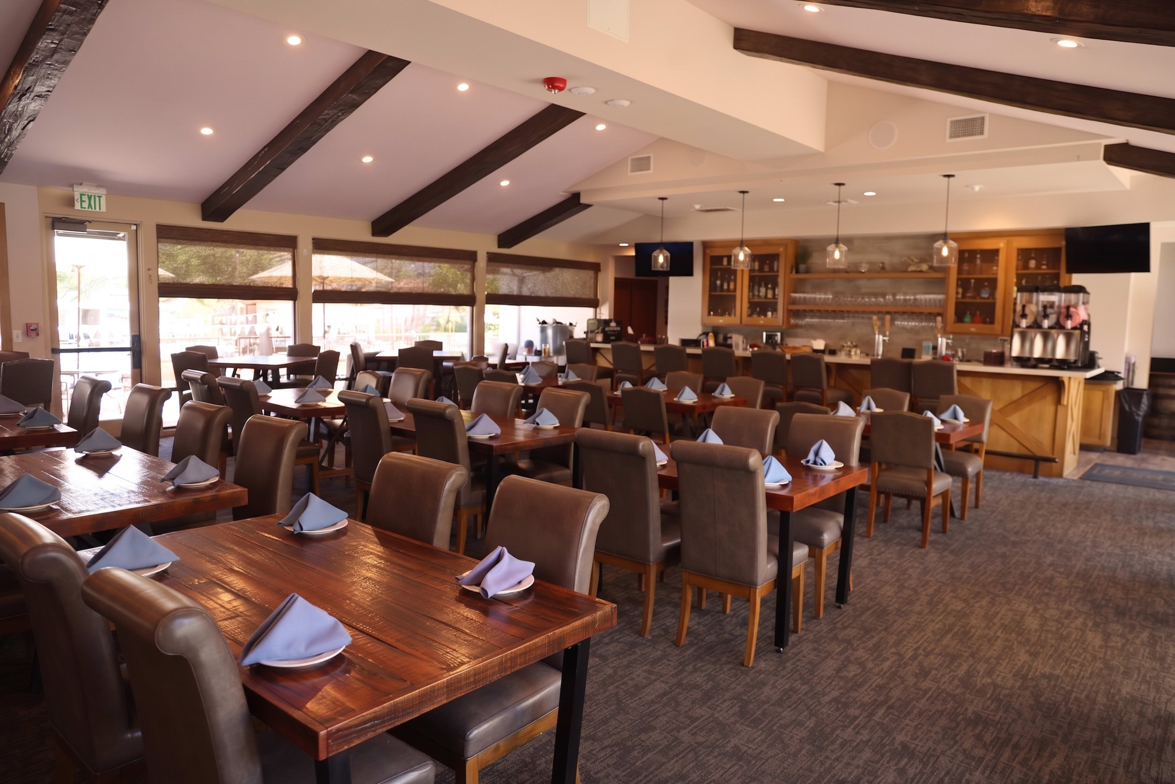 The image shows a spacious restaurant interior with wooden beams, neatly set tables, and a bar area in the background.