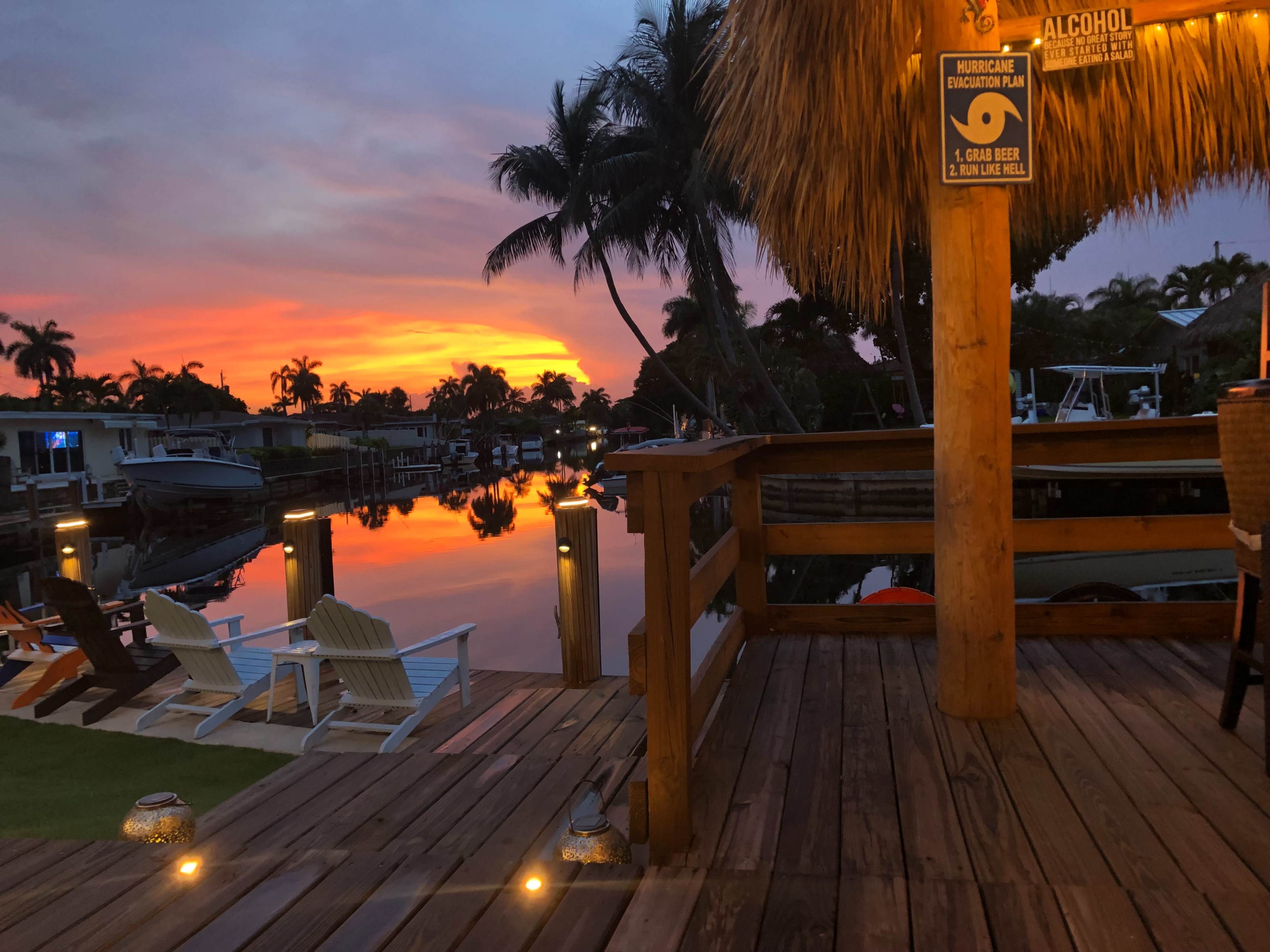 A wooden deck overlooks a calm waterway reflecting a vibrant sunset, with boats docked alongside and palm trees in the background.
