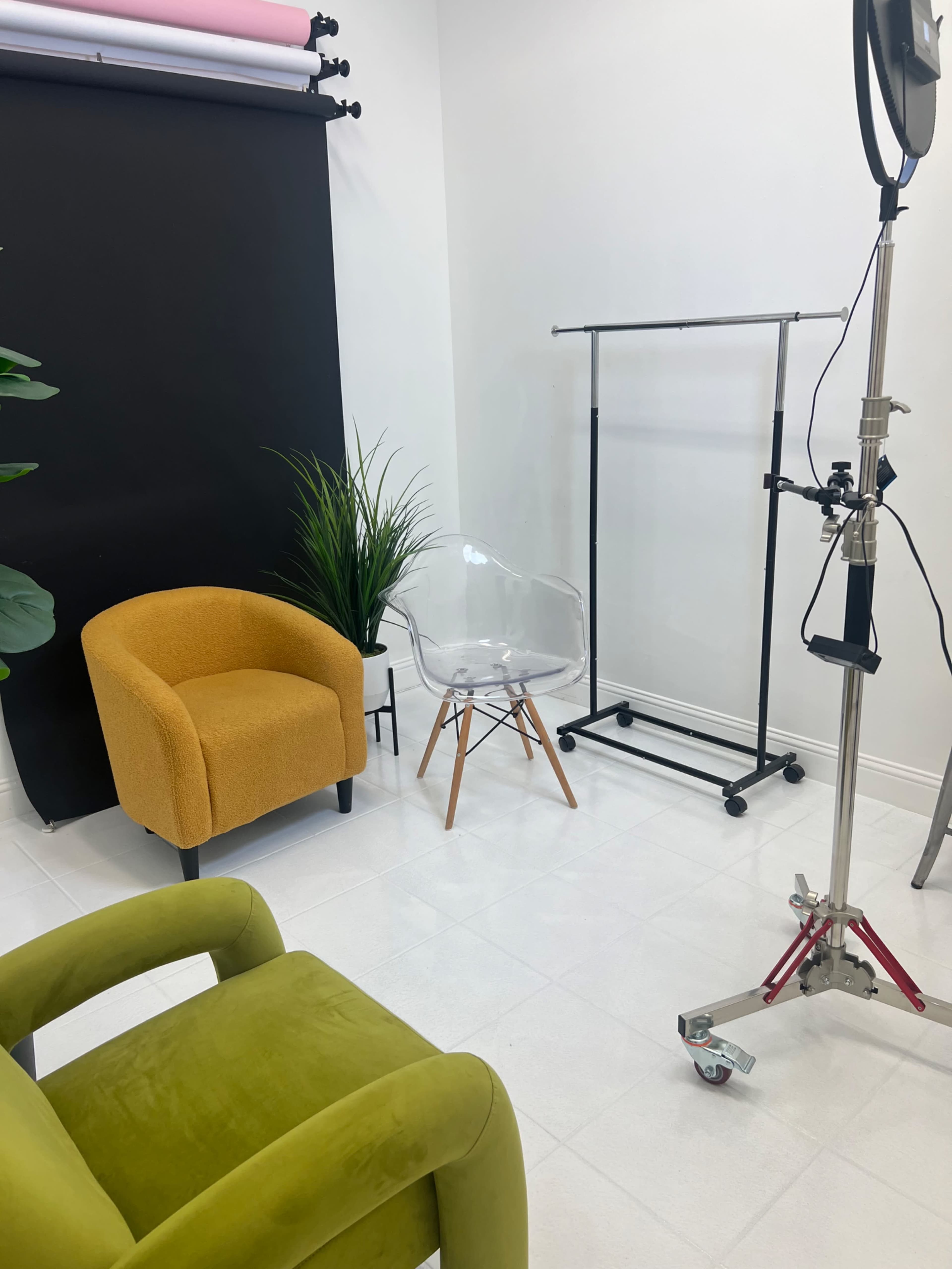 The image features a minimalistic studio setup with a bright yellow chair, a clear plastic chair, and a clothing rack against a black backdrop, accompanied by a light stand and a potted plant.