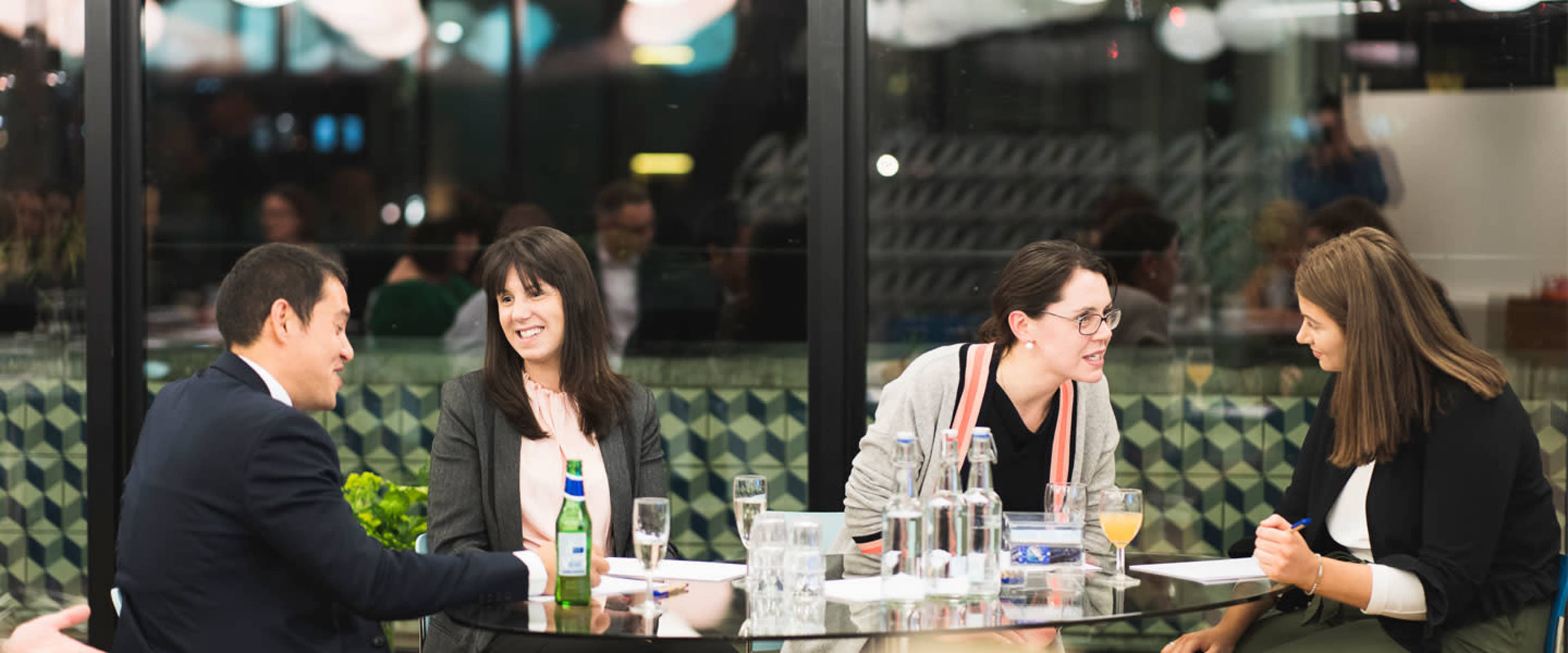 A group of five people sits around a glass table in a modern, well-lit setting, engaging in conversation.