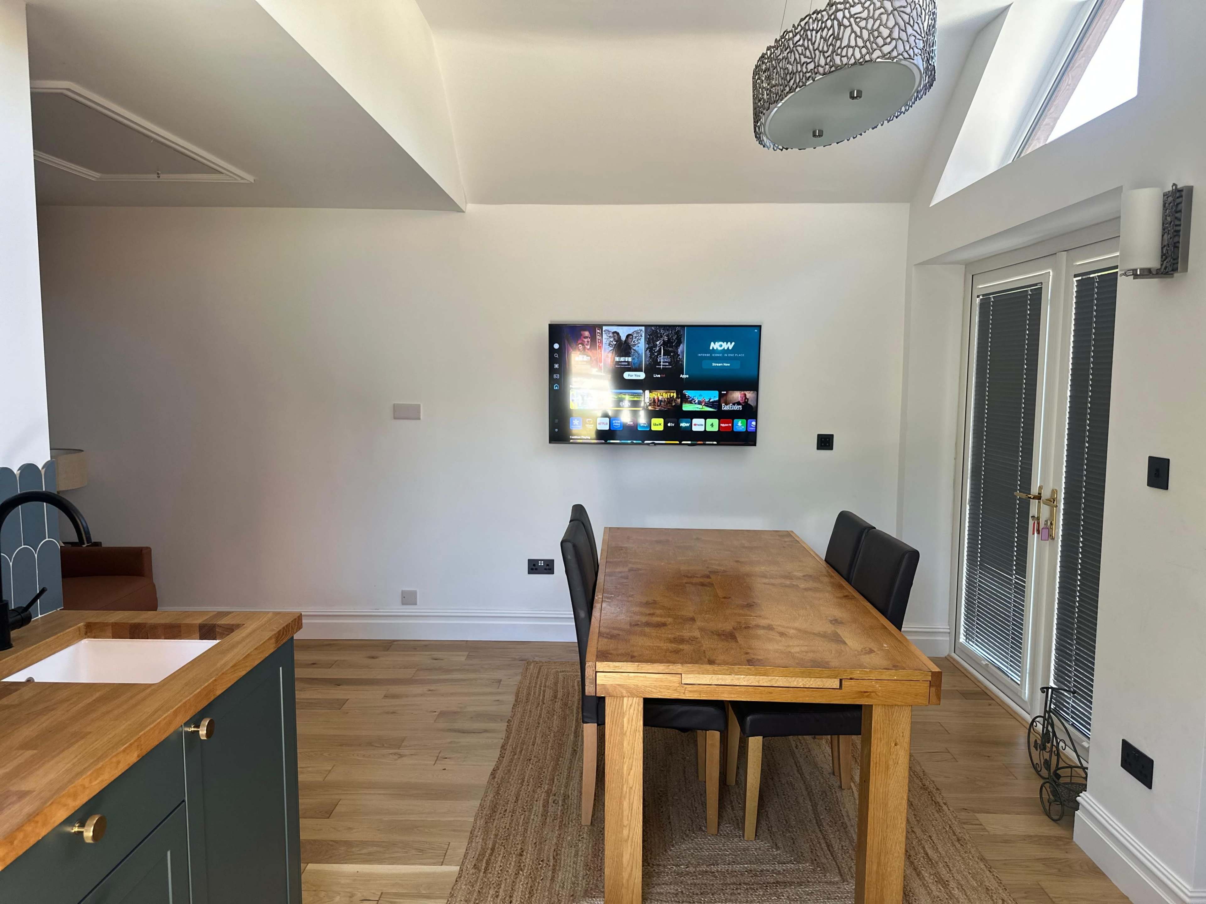 A wooden dining table with black chairs is positioned in a bright room featuring a wall-mounted television and large windows.