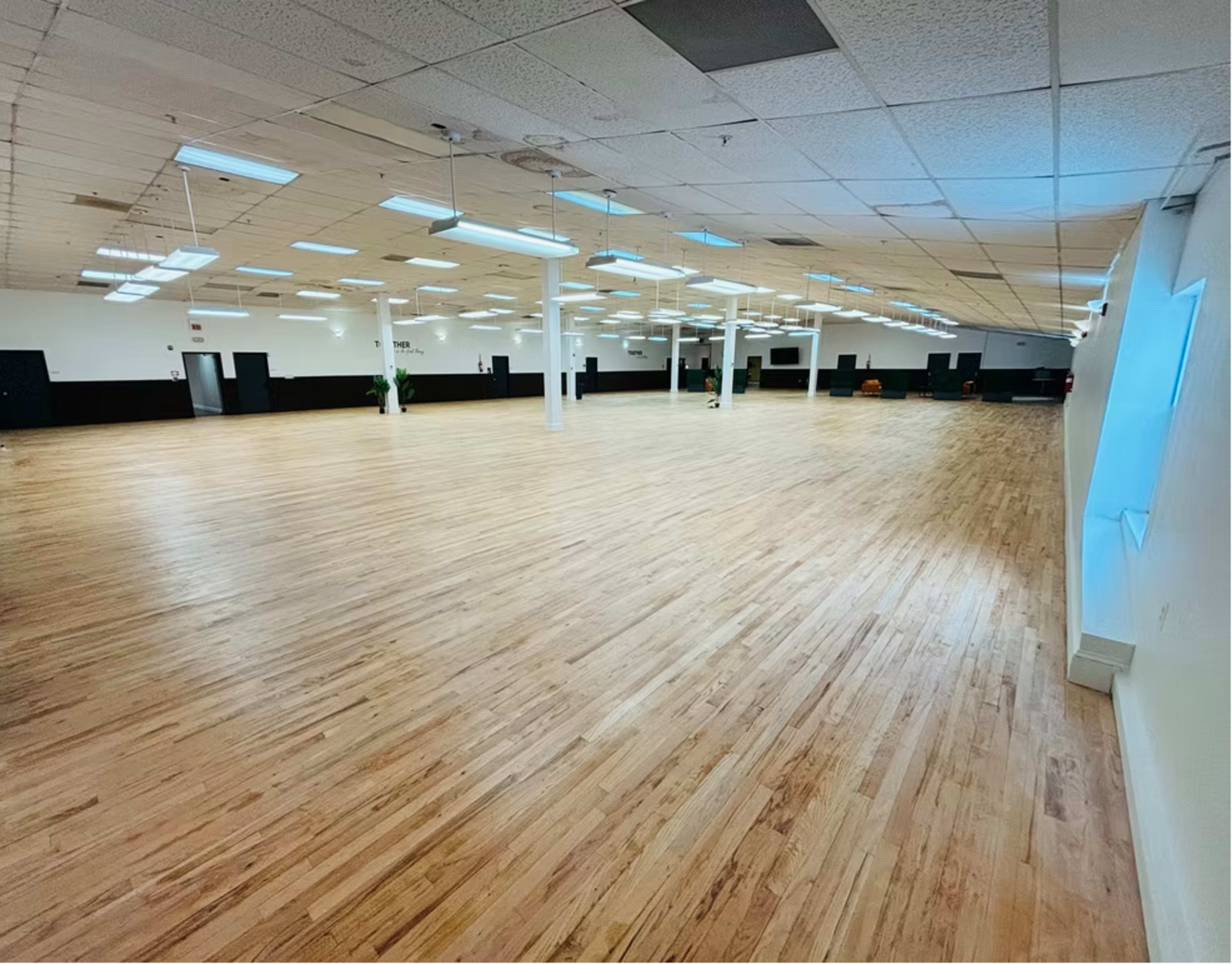 The image shows a large, empty room with wooden flooring, overhead lights, and minimal furnishings along the walls.