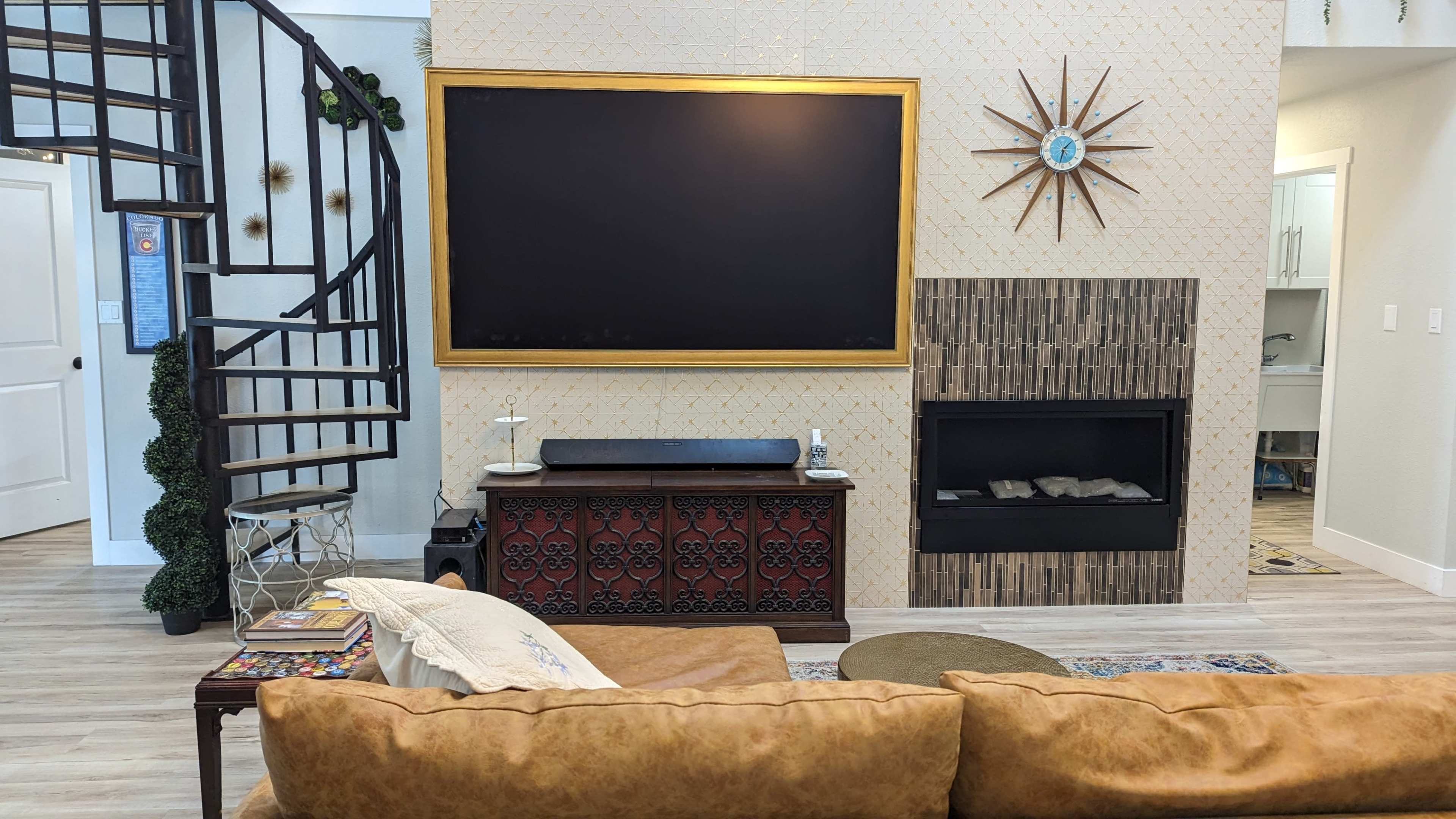 The living room features a spiral staircase, a large television mounted on a wall with decorative wallpaper, and a fireplace with a modern stone facade, all centered around a low sofa and coffee table.
