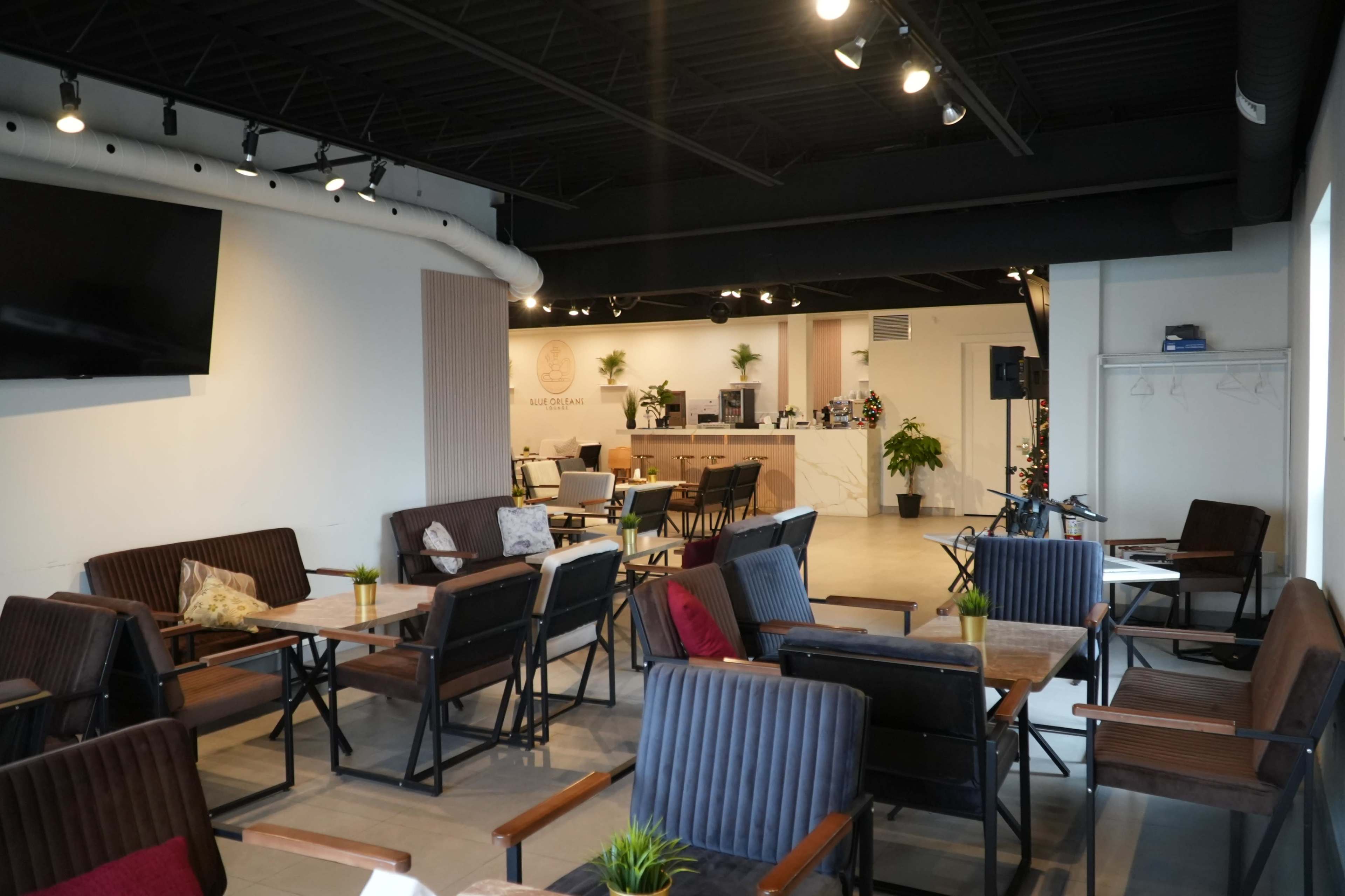 The image shows a modern café interior with rows of wooden seating and tables, a barista counter in the background, and decorative plants throughout the space.