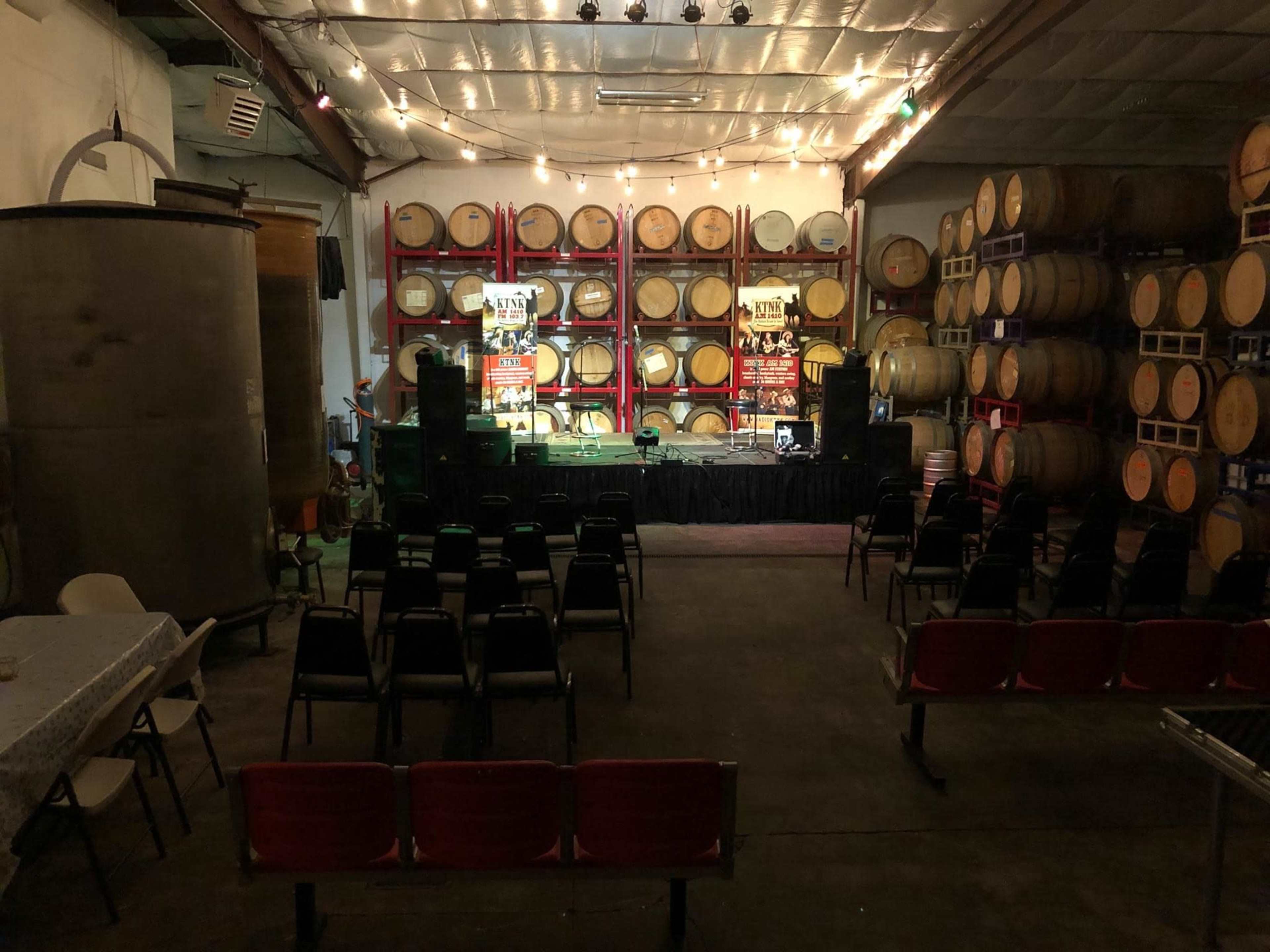 An event space featuring rows of chairs facing a stage, set against a backdrop of stacked wine barrels and ambient lighting.